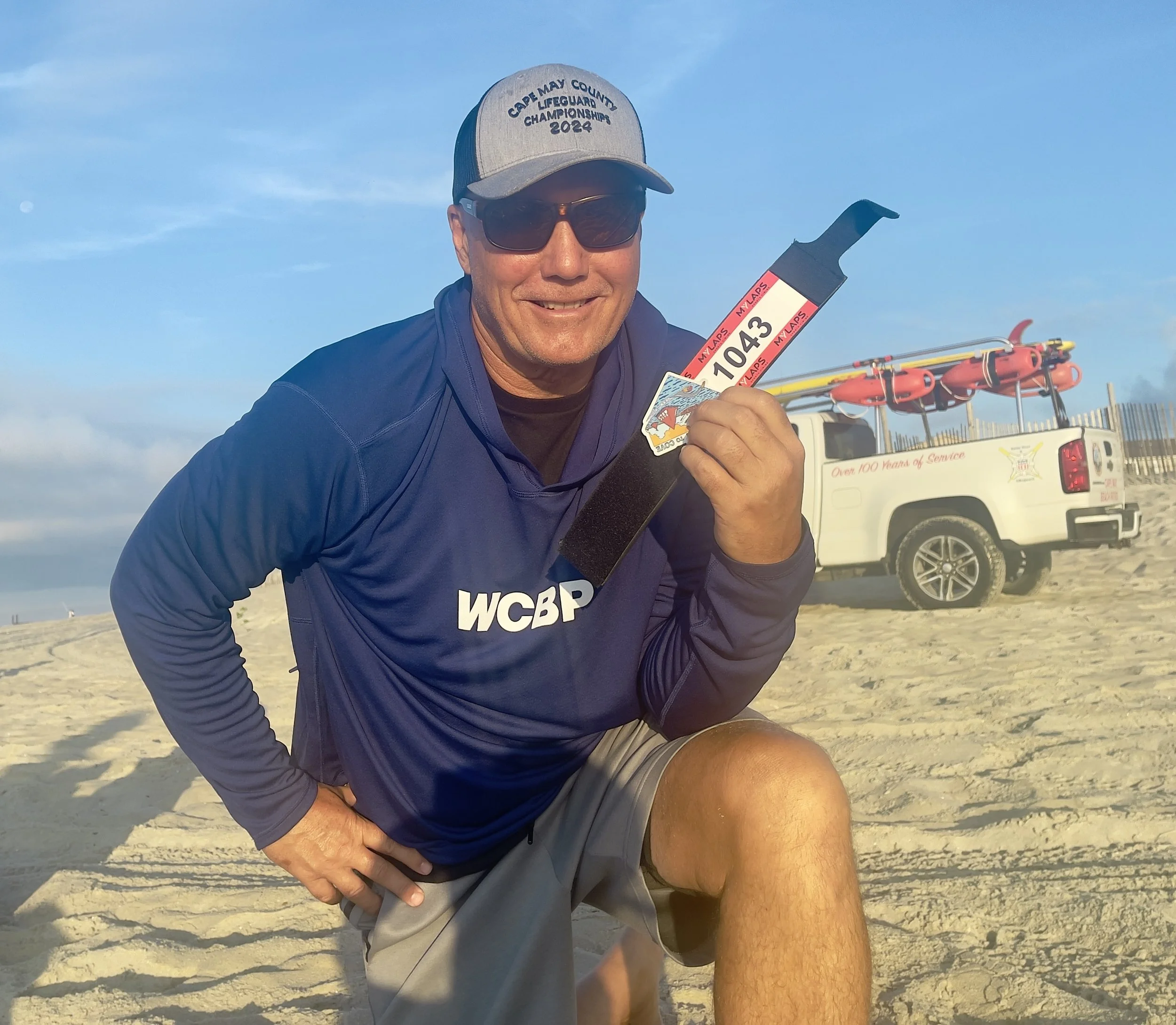 A smiling man at the beach wearing a cap, sunglasses, and a WCWP shirt, holding a surfboard leash with race numbers, and standing next to another person who is crouching. In the background, there are orange rescue tubes on a vehicle and a clear sky.