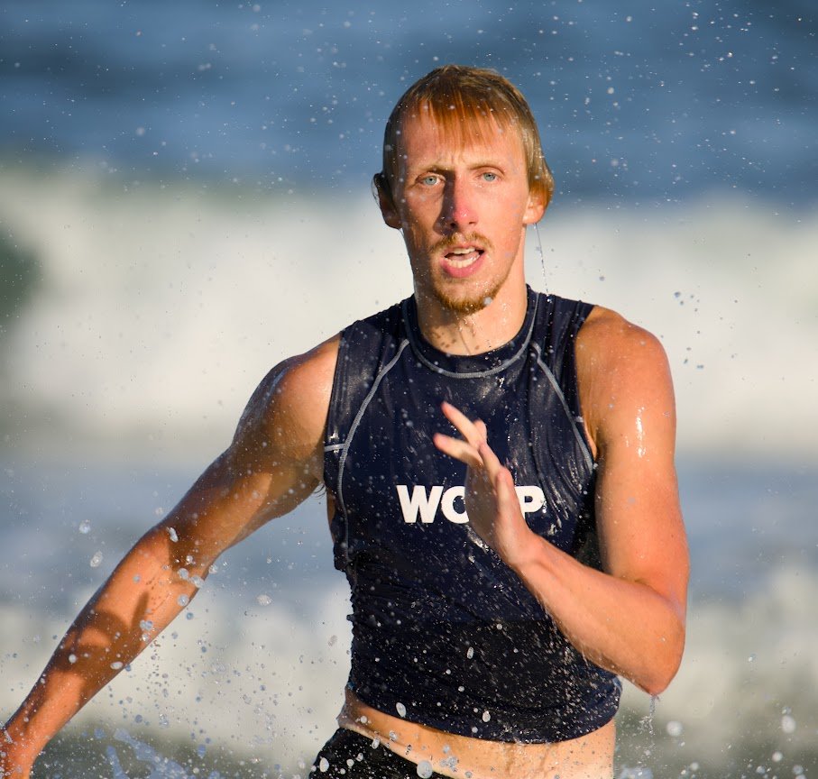 A man running through the ocean waves during a race or workout at the beach, wearing a black sleeveless athletic top.