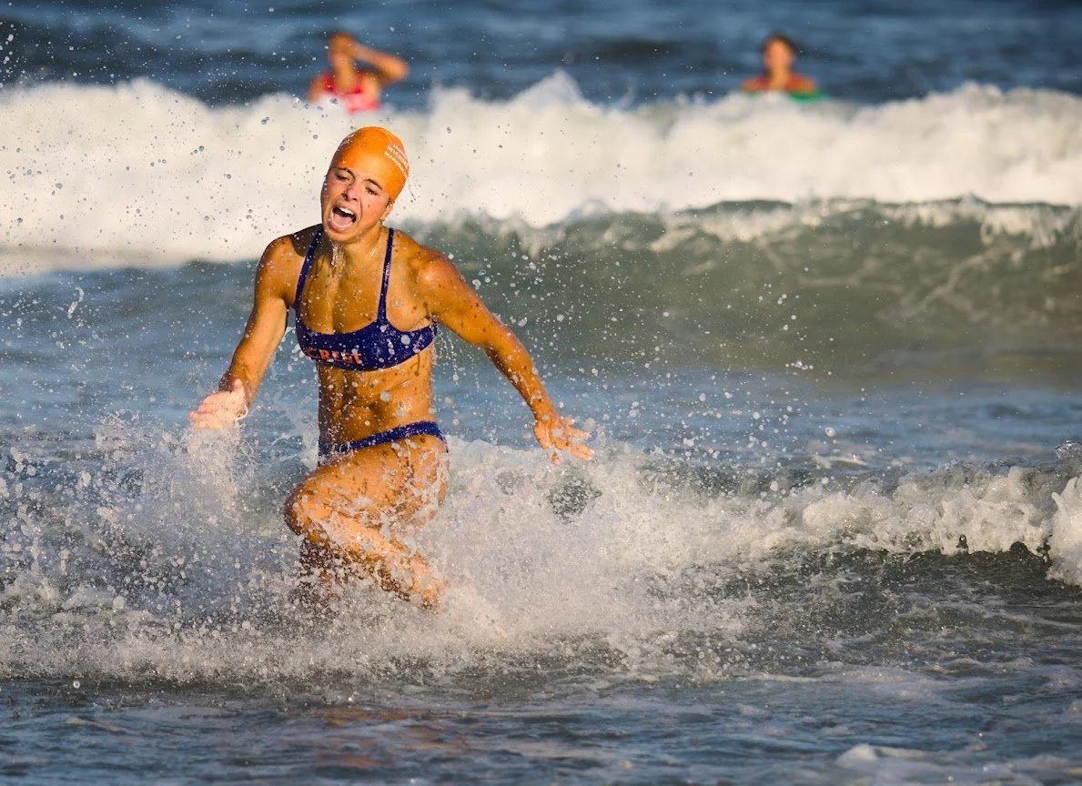 A woman in a blue swimsuit and orange swim cap is running through the ocean waves, appearing to be playing or competing in a water sport, with two other swimmers visible in the background.