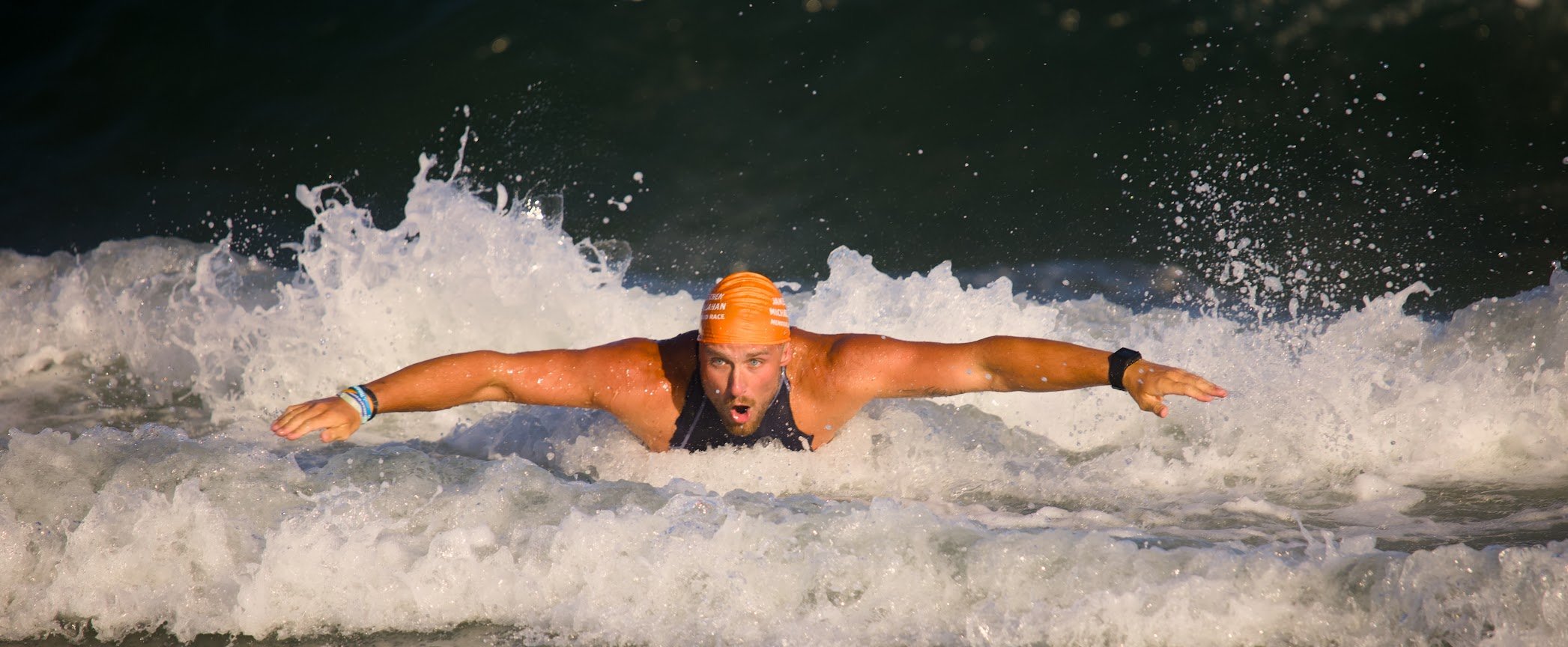 A male swimmer wearing an orange swim cap and black swimsuit swimming front crawl in open water, creating a splash as he propels forward.