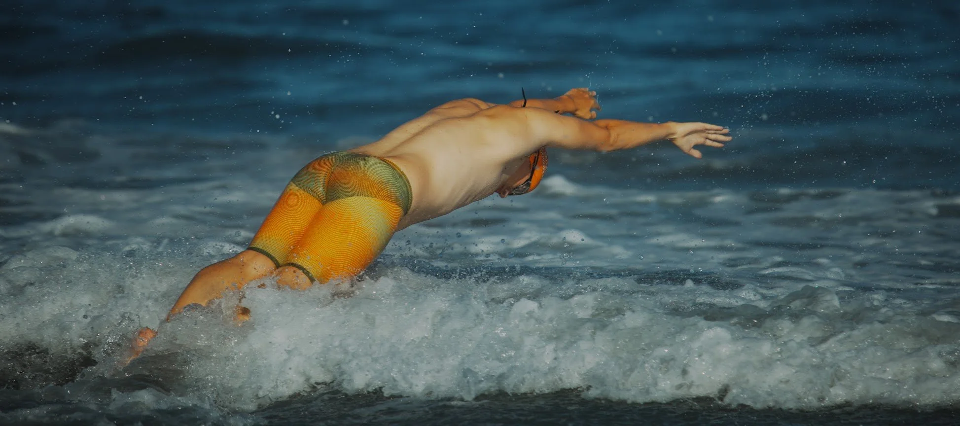 A person swimming in the ocean wearing bright orange and green swimwear, reaching forward while diving through the water.