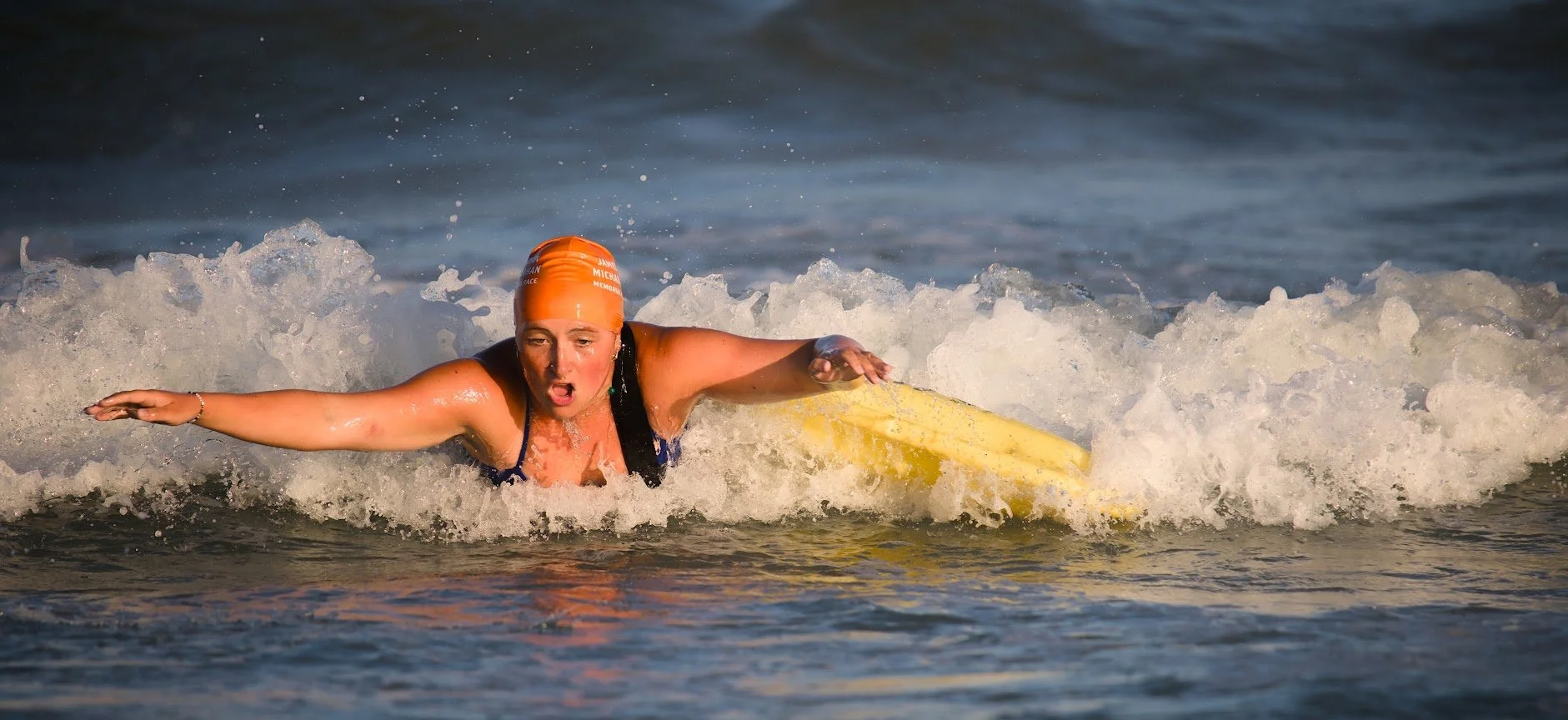 Female swimmer in orange swim cap and blue swimsuit paddling on a yellow surfboard through ocean waves.