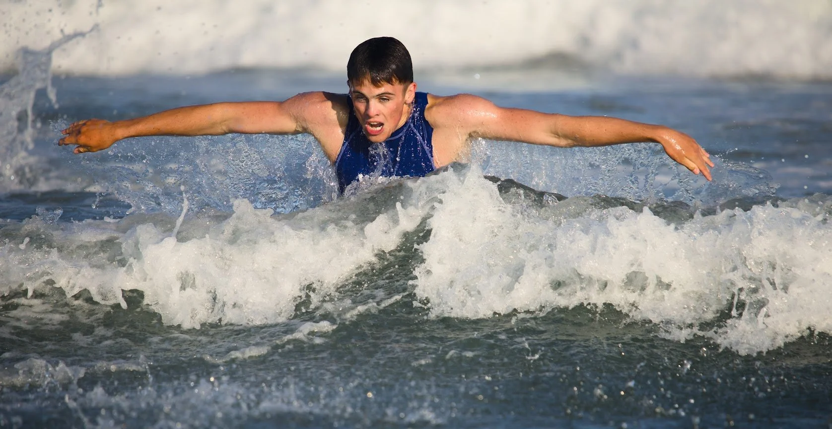 A young boy in a blue swimsuit paddling through ocean waves, with a determined expression on his face.