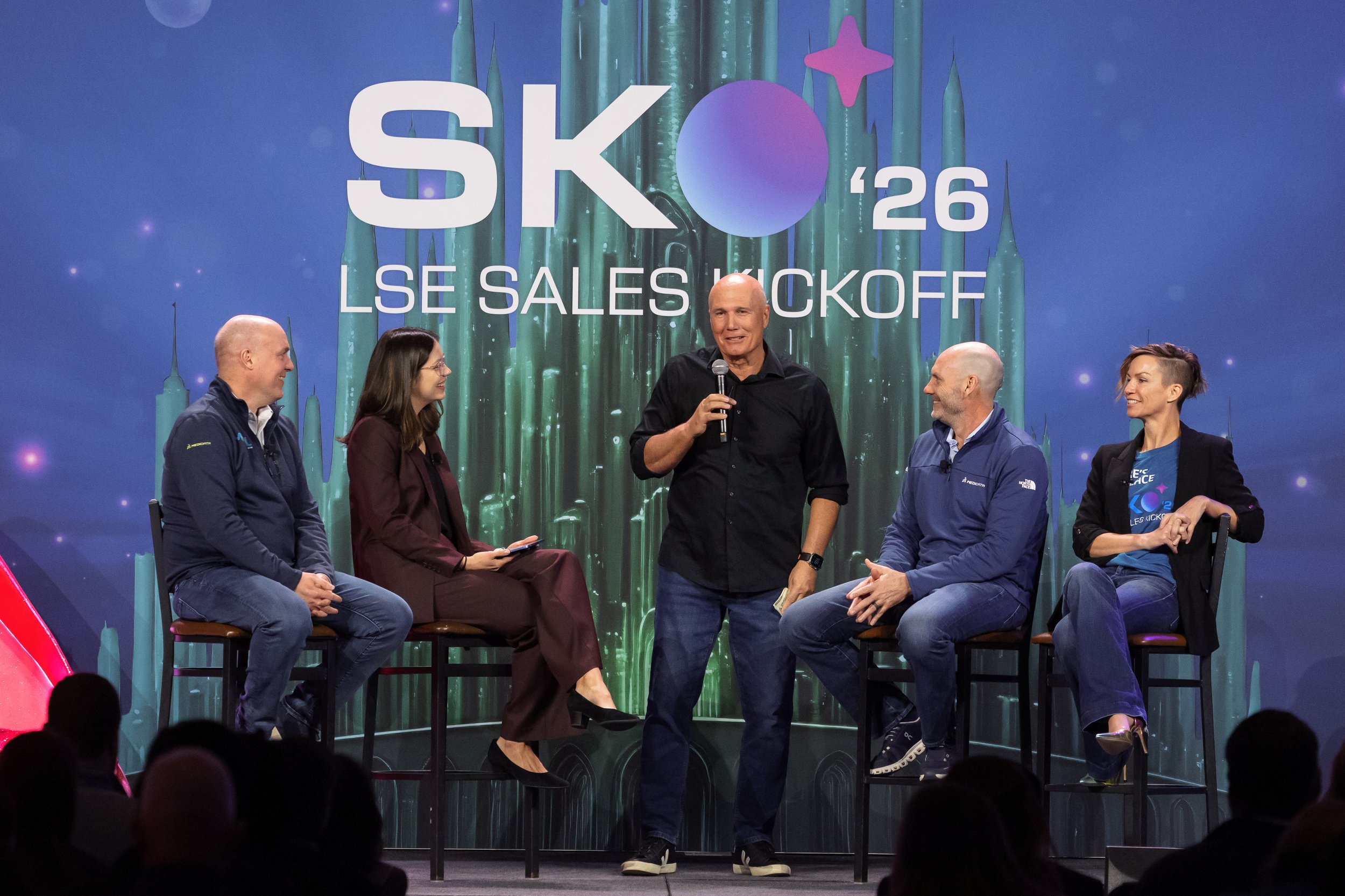 Six people seated and one standing on stage during an event titled 'SKO '26 LSE Sales Kickoff'. The background displays a large screen with the event name and a futuristic design with green pillars and purple accents. The participants are engaged in conversation, some holding microphones.