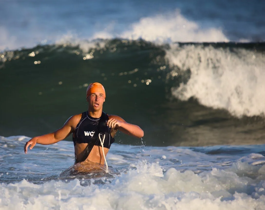 A female athlete wearing a black triathlon suit and orange swim cap exiting the ocean after swimming.
