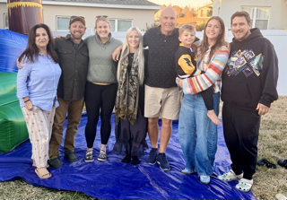 Group of eight people standing outdoors on a blue tarp in front of a house, smiling and posing for the photo.