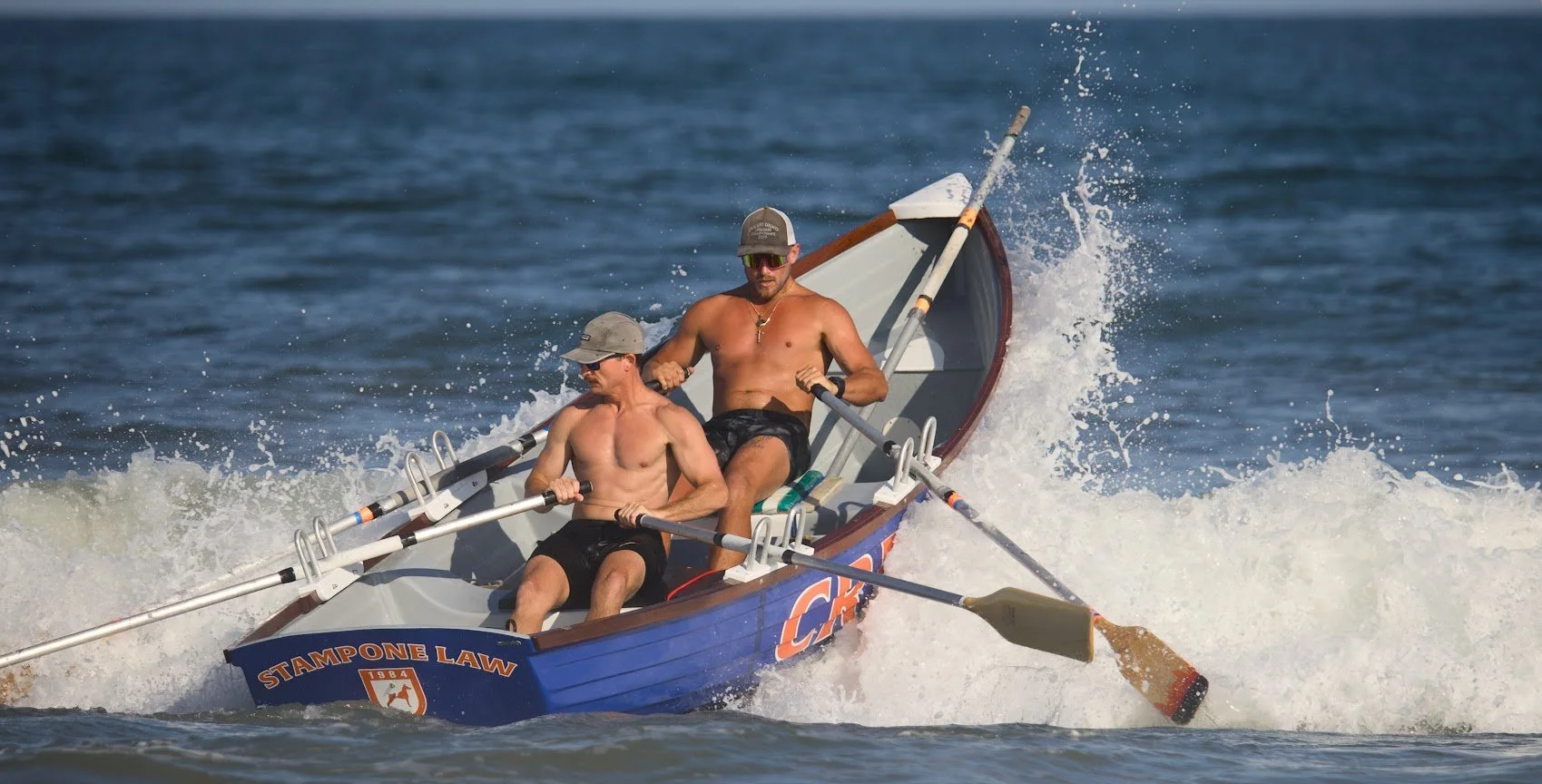 Two shirtless men wearing caps and sunglasses rowing a small blue and white boat on the water, creating a splash.
