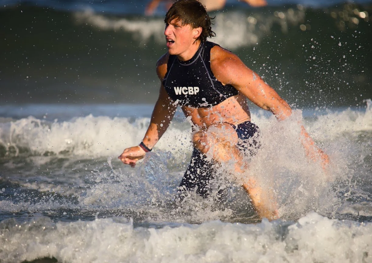 A young man in a black wetsuit with 'WCBP' on it is crouching in the ocean waves, struggling to stand or move forward amid splashing water.