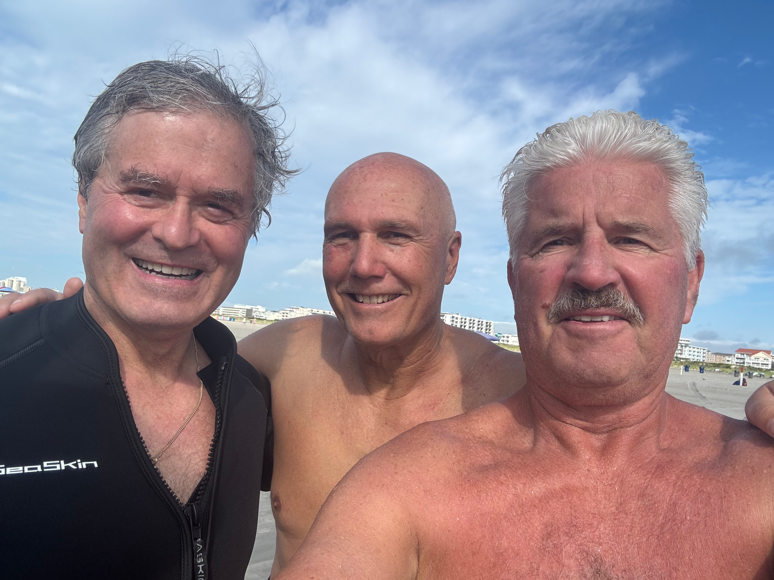 Three men are smiling and posing for a photo on the beach, with buildings and a blue sky with some clouds in the background.