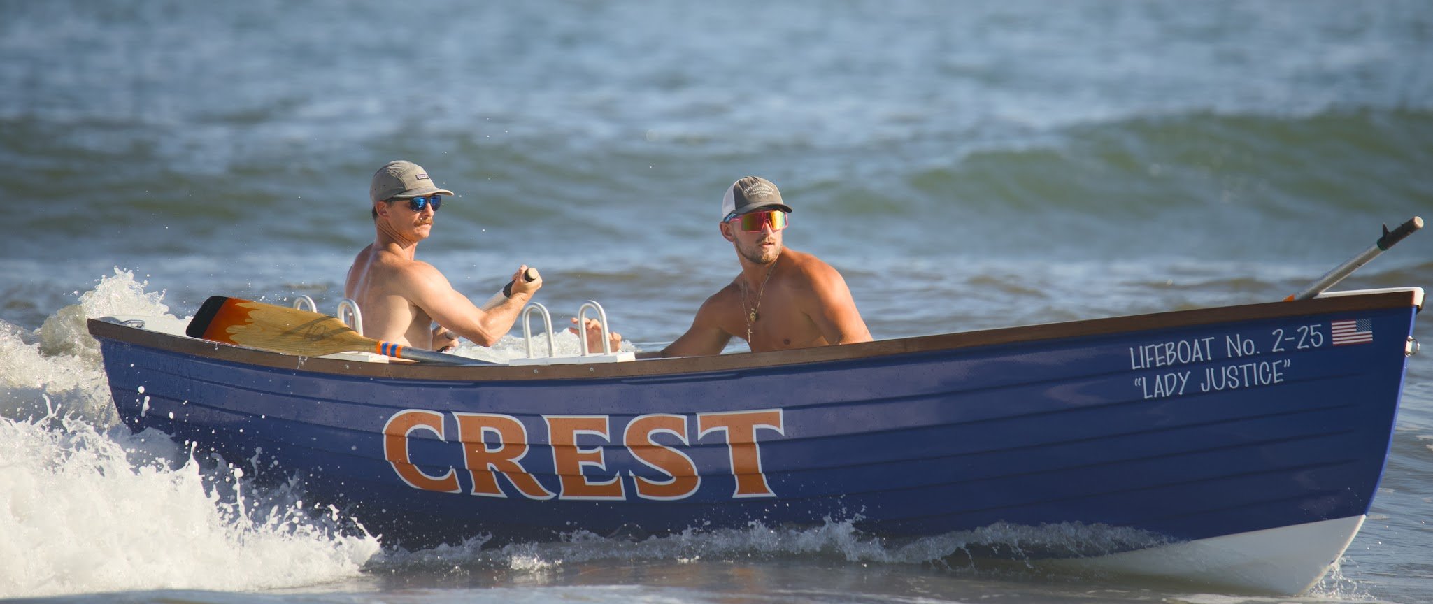 Two shirtless men with caps and sunglasses riding in a small blue boat on the water. The boat has the words 'Crest' and 'Lady Justice' written on it.