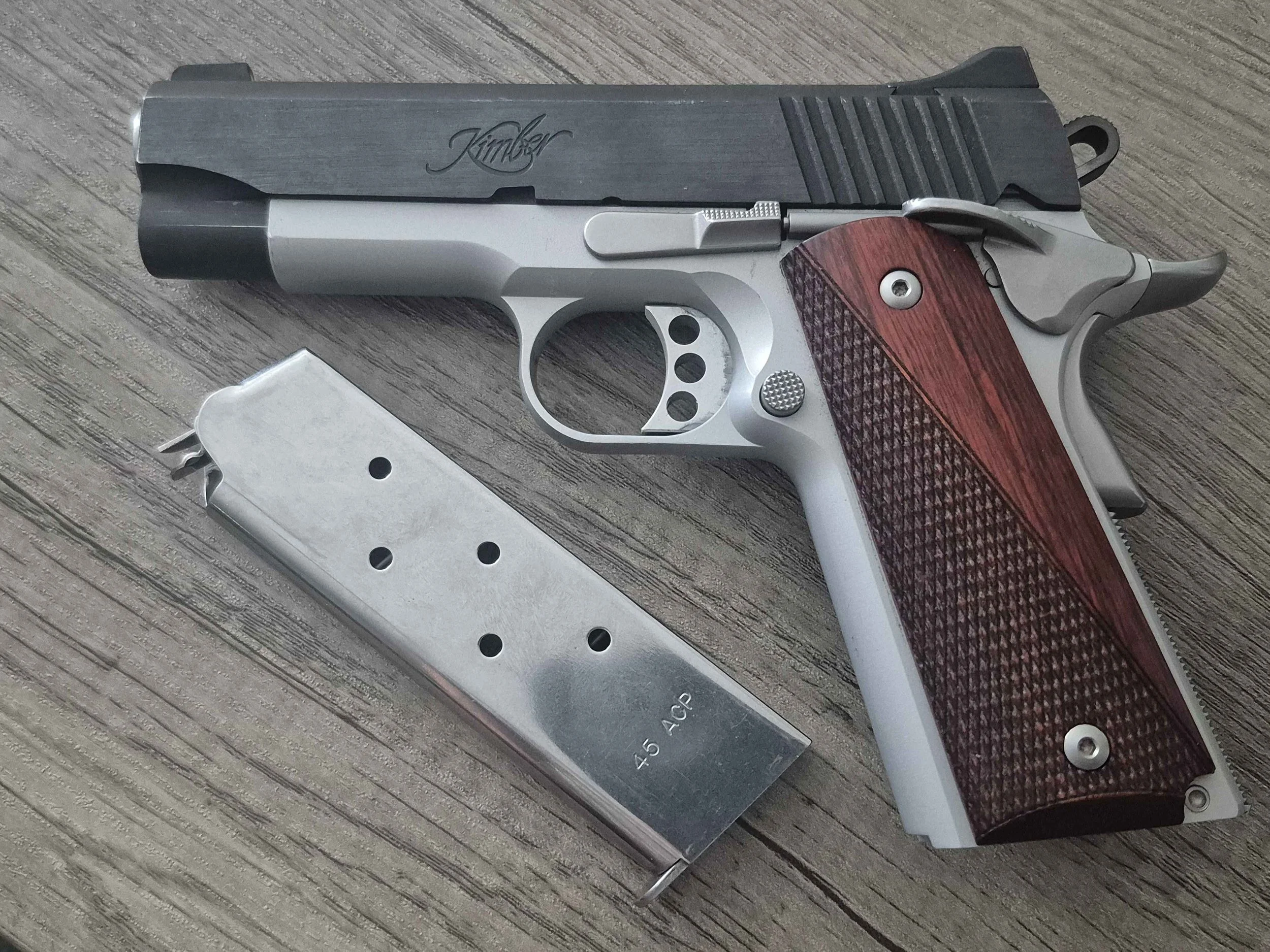 A semi-automatic Kimber handgun with a stainless steel slide and a red checkered wooden grip, placed on a wooden surface. The slide has the Kimber logo. A magazine is partially visible below the gun.