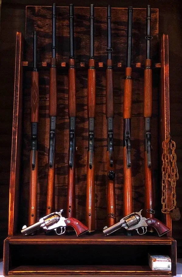 Display case with seven rifles arranged vertically, two revolvers placed at the bottom, and a box of ammunition on the bottom right.
