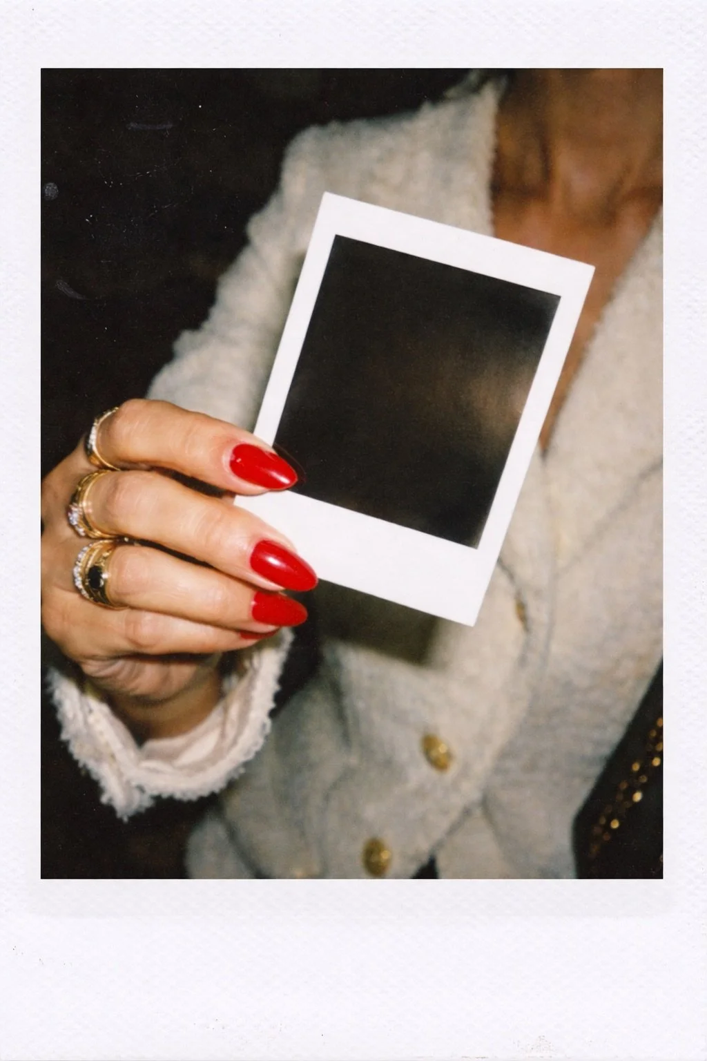 A woman holding an empty instant photo with her nails painted red, wearing multiple rings, and a white textured coat with gold buttons.