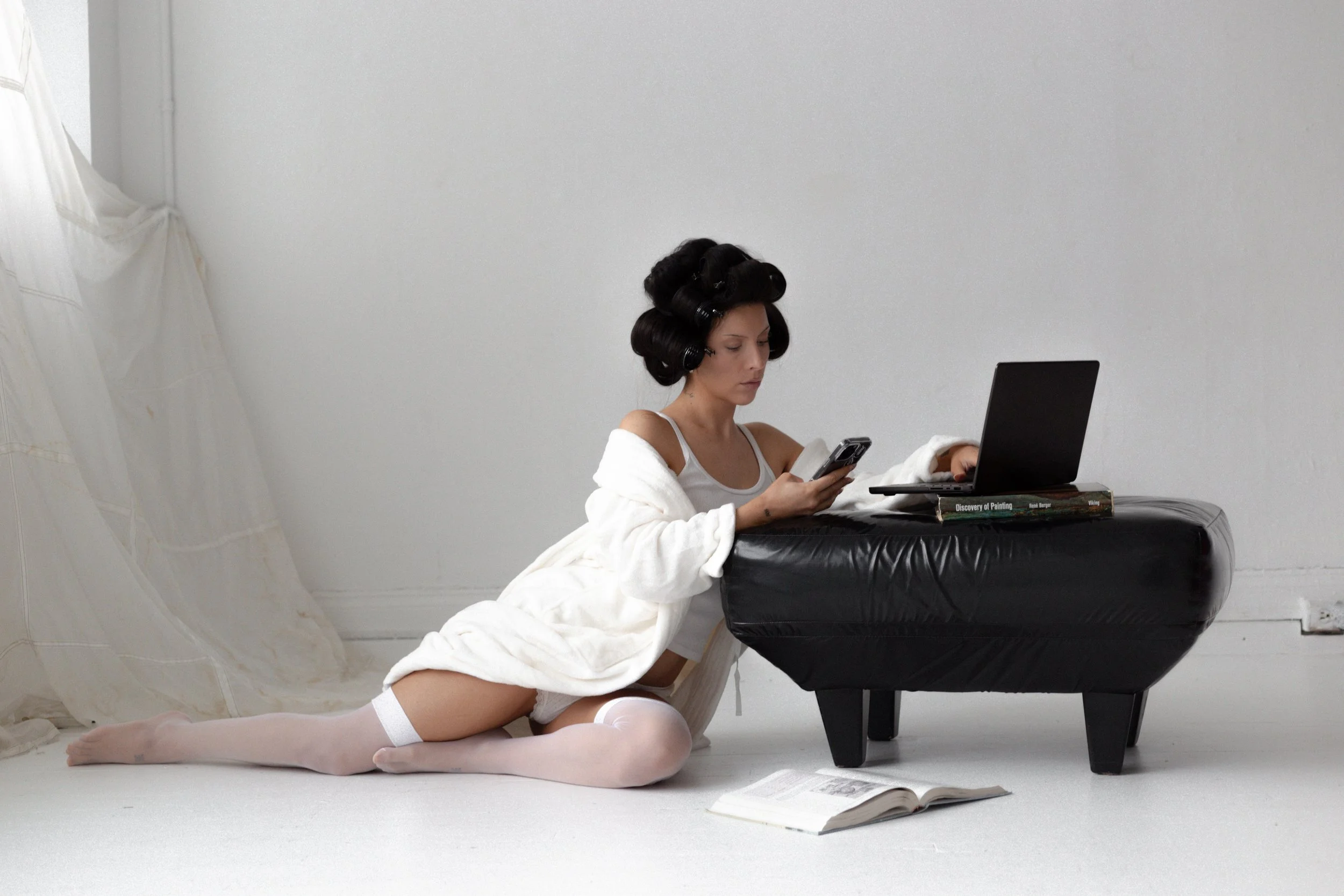 Young woman with rollers in her hair sitting on the floor, wearing white lingerie and knee-high socks, using a smartphone. There is an open book on the floor, a black piano, and a white blanket or towel around her shoulders. The setting is minimalistic with a plain white background.