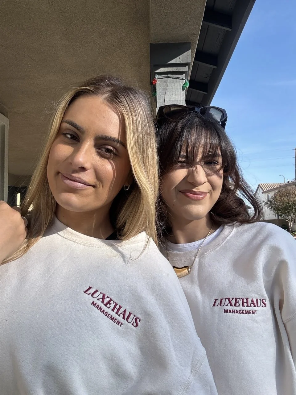 Two women in white sweatshirts with 'LUXEHAUS MANAGEMENT' embroidered on, taking a selfie outdoors on a sunny day.