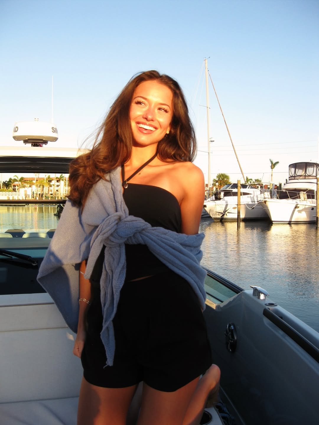 A woman with long brown hair smiling on a boat docked at a marina with yachts and boats, in warm sunlight, wearing a black strapless top, shorts, and a gray sweater tied around her shoulders.