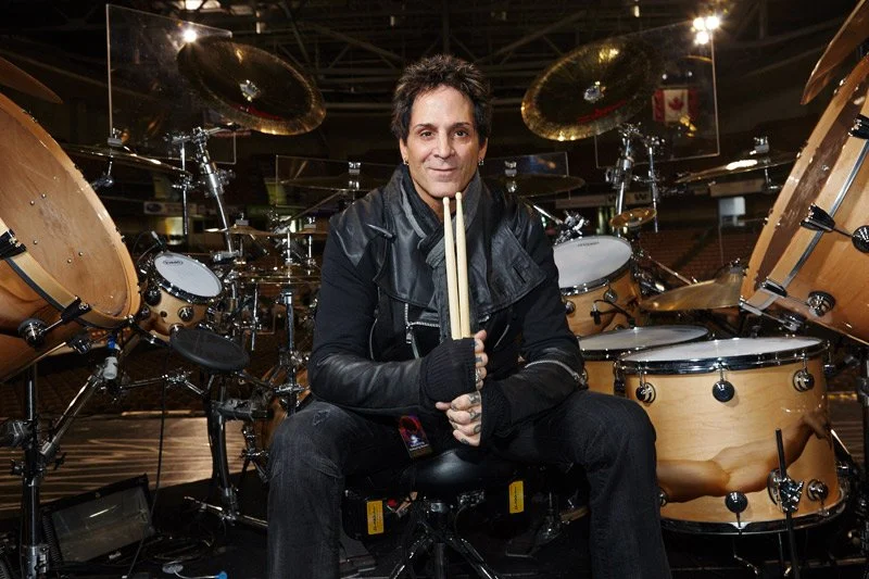 A man sitting in front of a drum set, holding drumsticks, with a smile, in a music studio or stage setting.