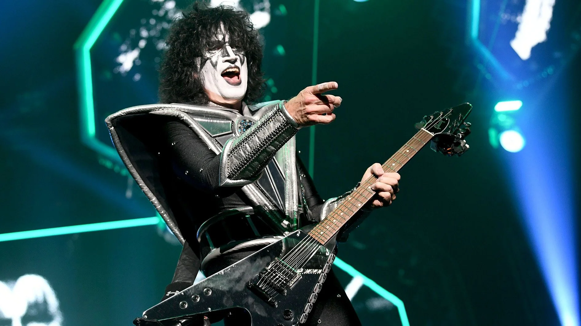 A man with face paint resembling KISS's Paul Stanley, wearing a metallic silver and black costume, playing an electric guitar on stage with green and blue stage lighting.