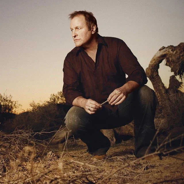 A man crouches outdoors at sunset, wearing a dark shirt and jeans, sitting beside a fallen tree.