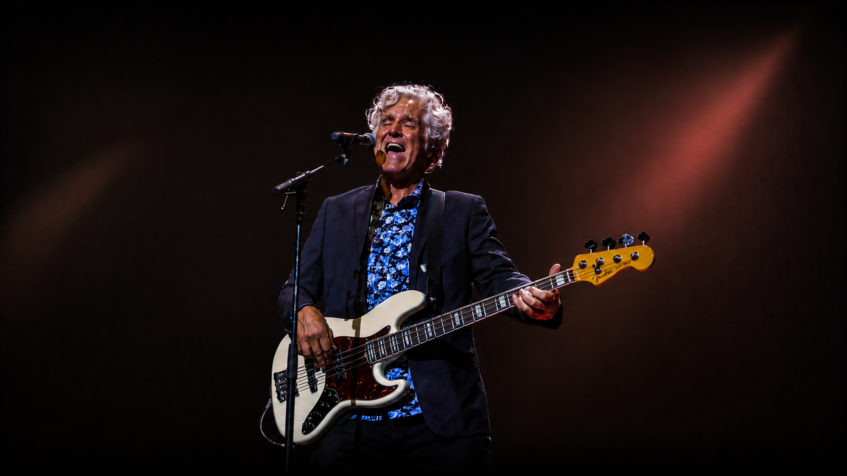 A male musician with curly gray hair singing passionately into a microphone while playing a bass guitar on stage, illuminated by stage lights.