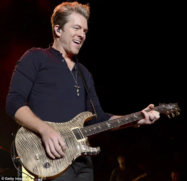 Male musician on stage playing an electric guitar, smiling, wearing a black shirt, with a microphone headset, in a dark setting.