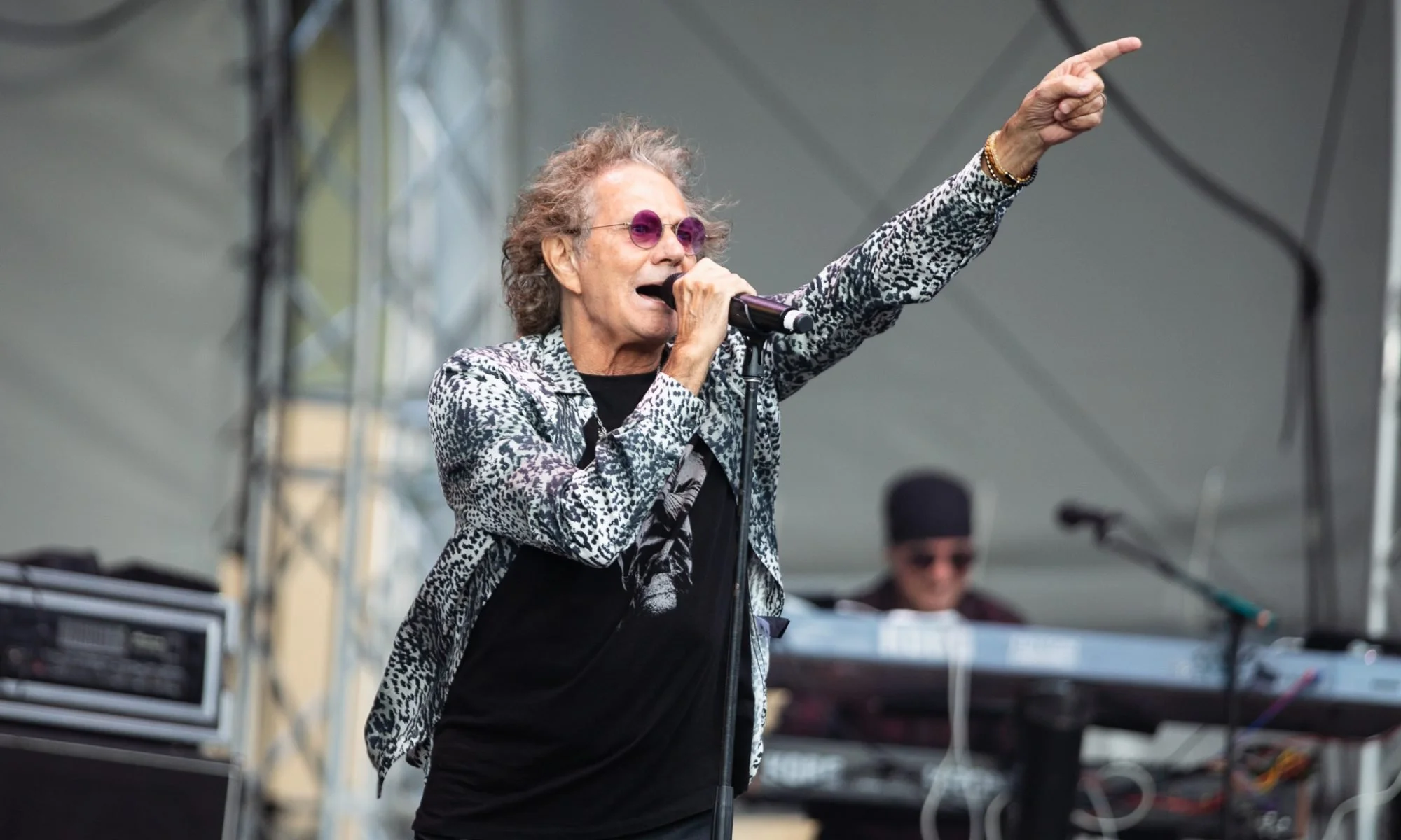 A woman with curly gray hair and purple sunglasses singing into a microphone on stage, wearing a leopard print jacket and pointing upward with her right hand. A musician with sunglasses is in the background.