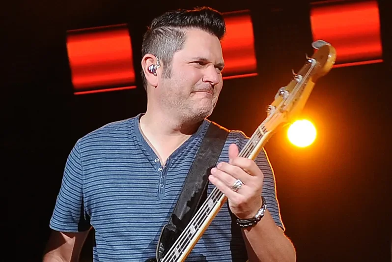 A man playing an electric bass guitar on stage, wearing a blue striped shirt and smiling with eyes closed, backlit by orange and red stage lights.