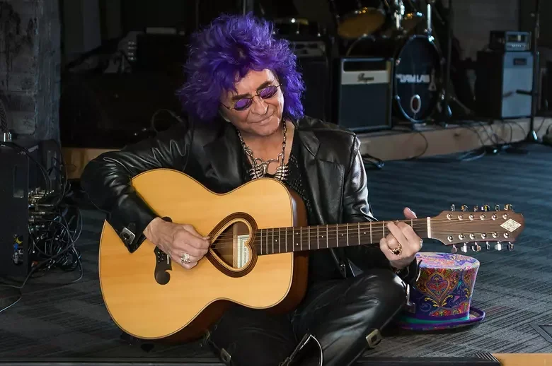 Person with purple curly hair and sunglasses sitting on the floor playing an acoustic guitar in a music studio or practice space.