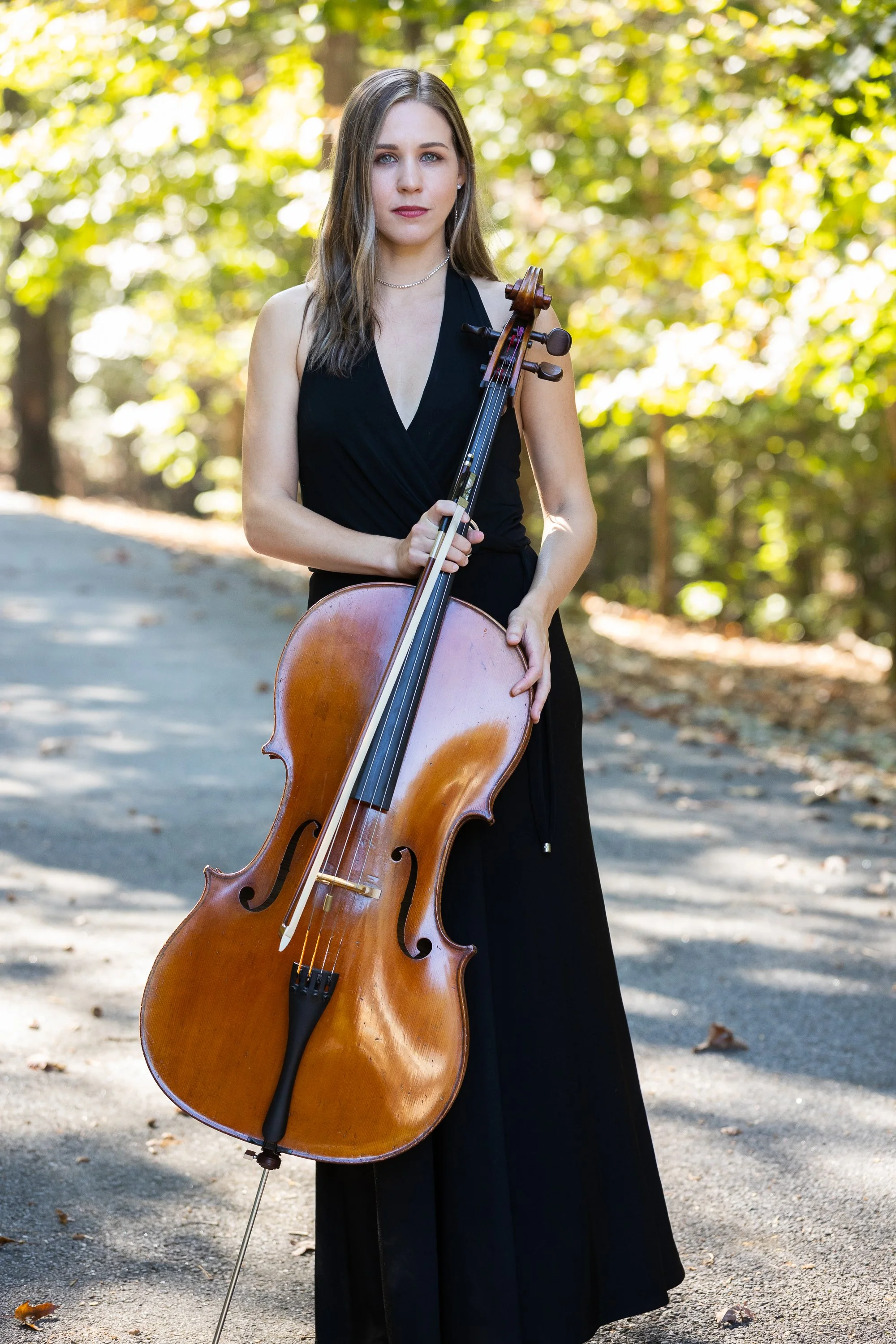 A woman with long brown hair wearing a black sleeveless dress, holding a cello with both hands, standing outdoors on a sunny day with trees and fallen leaves in the background.