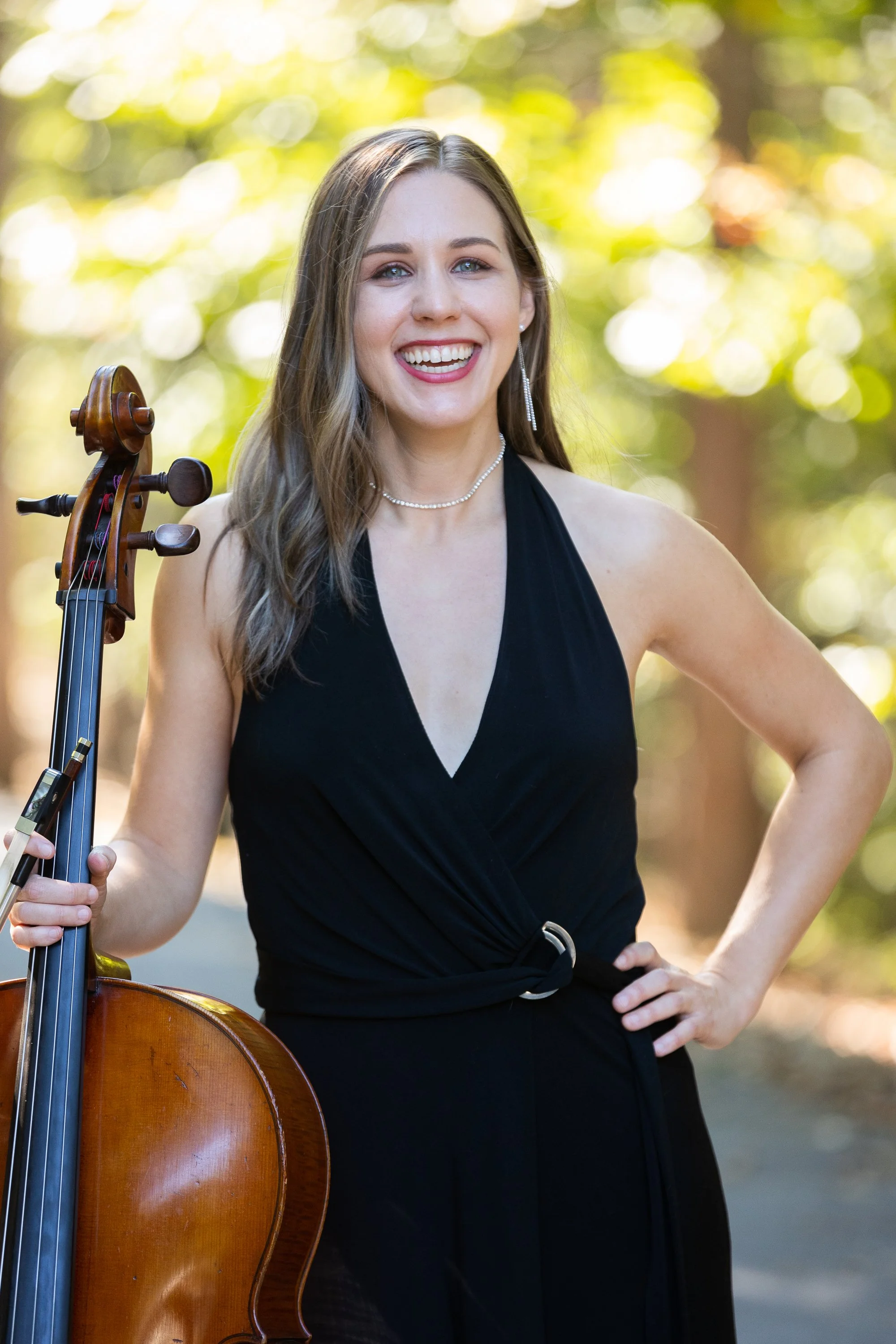 A woman with long brown hair and blue eyes smiling and holding a cello outdoors, wearing a black dress with a halter neckline, in front of a background of blurred trees and sunlight.