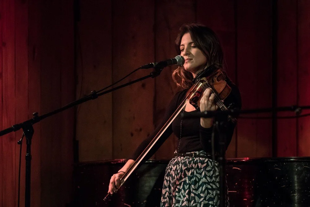 A woman on stage playing the violin with a microphone, eyes closed, under warm lighting with a wooden background.