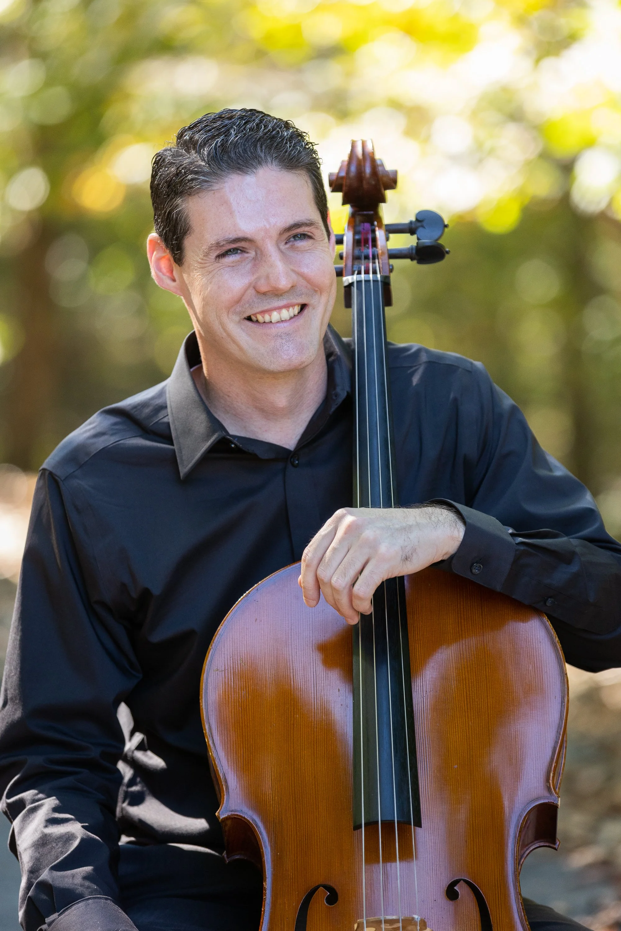 A man with short dark hair smiling and holding a cello outdoors with a blurred background of trees and sunlight.