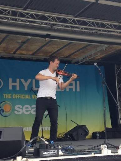 A young man playing a violin on an outdoor stage during a performance, with a large blue banner in the background.
