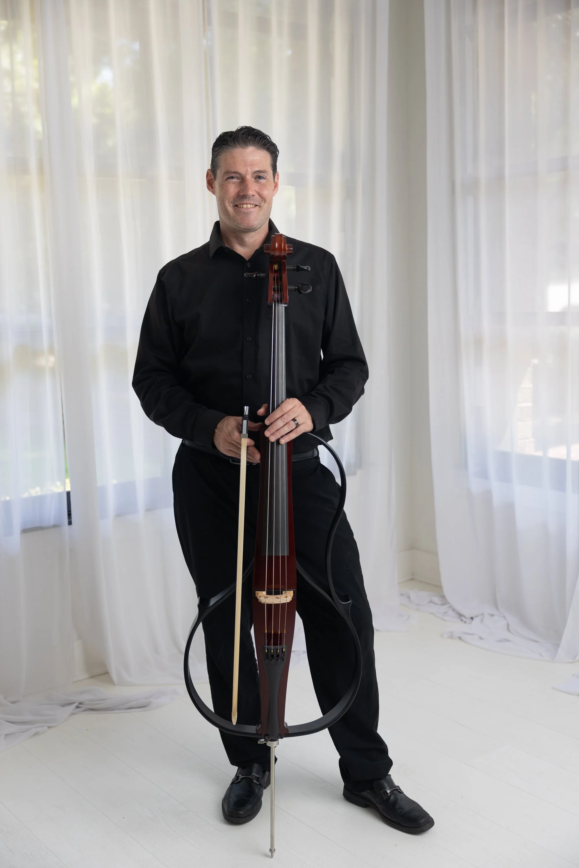 A man with short dark hair smiling and holding a cello outdoors with a blurred background of trees and sunlight.