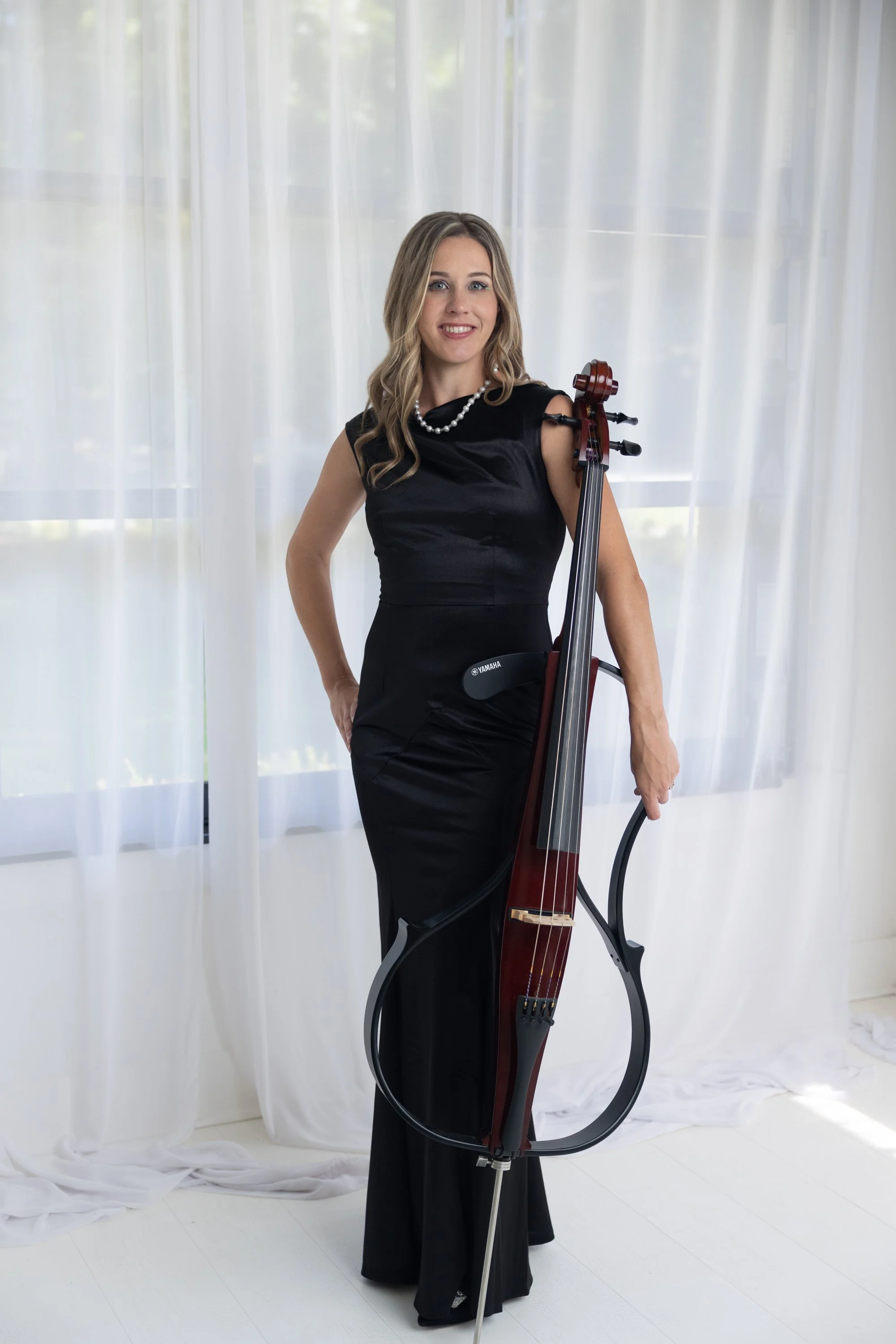 A woman with long brown hair and blue eyes smiling and holding a cello outdoors, wearing a black dress with a halter neckline, in front of a background of blurred trees and sunlight.
