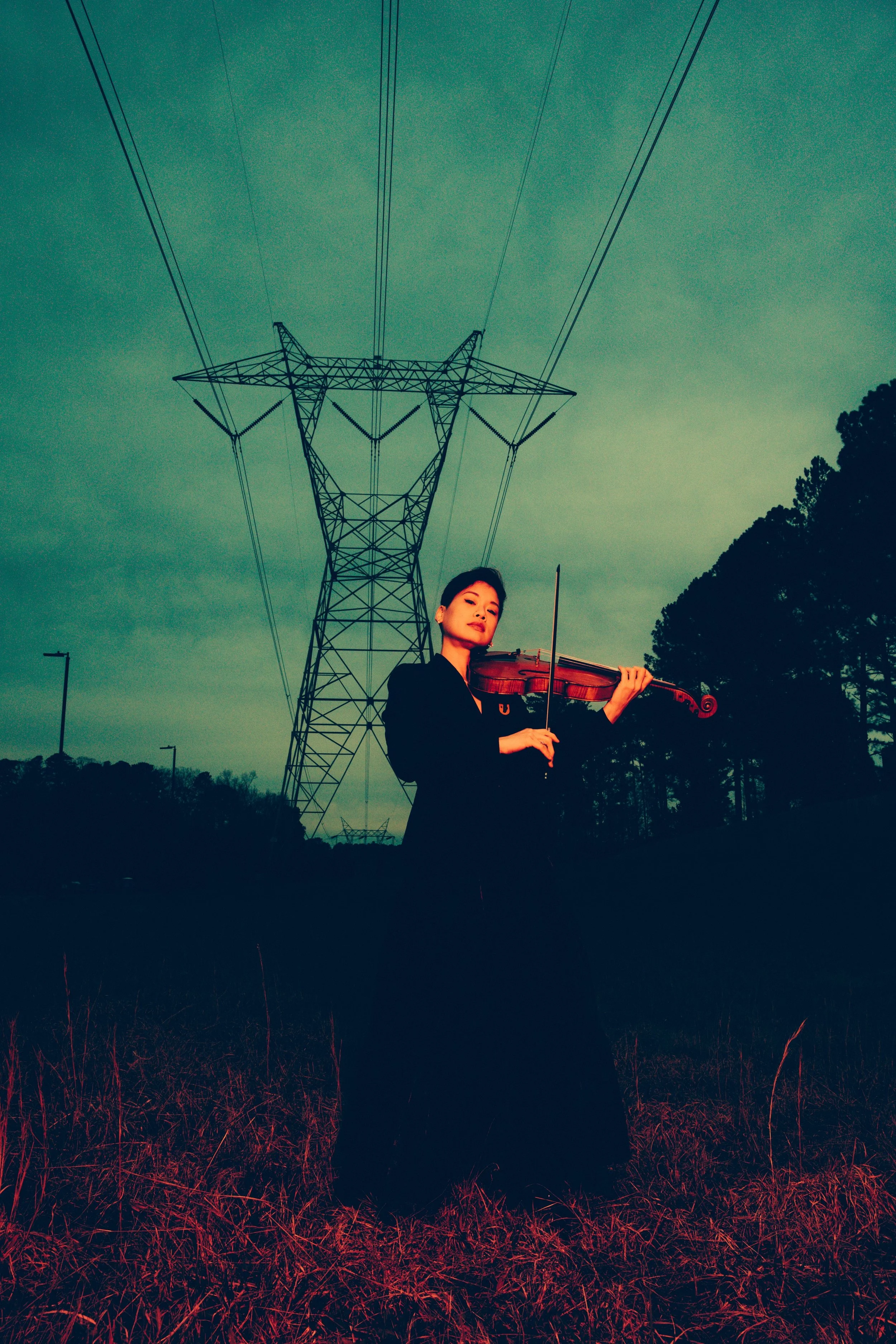 A woman playing the violin outdoors near power lines and trees at dusk or dawn with a greenish sky.