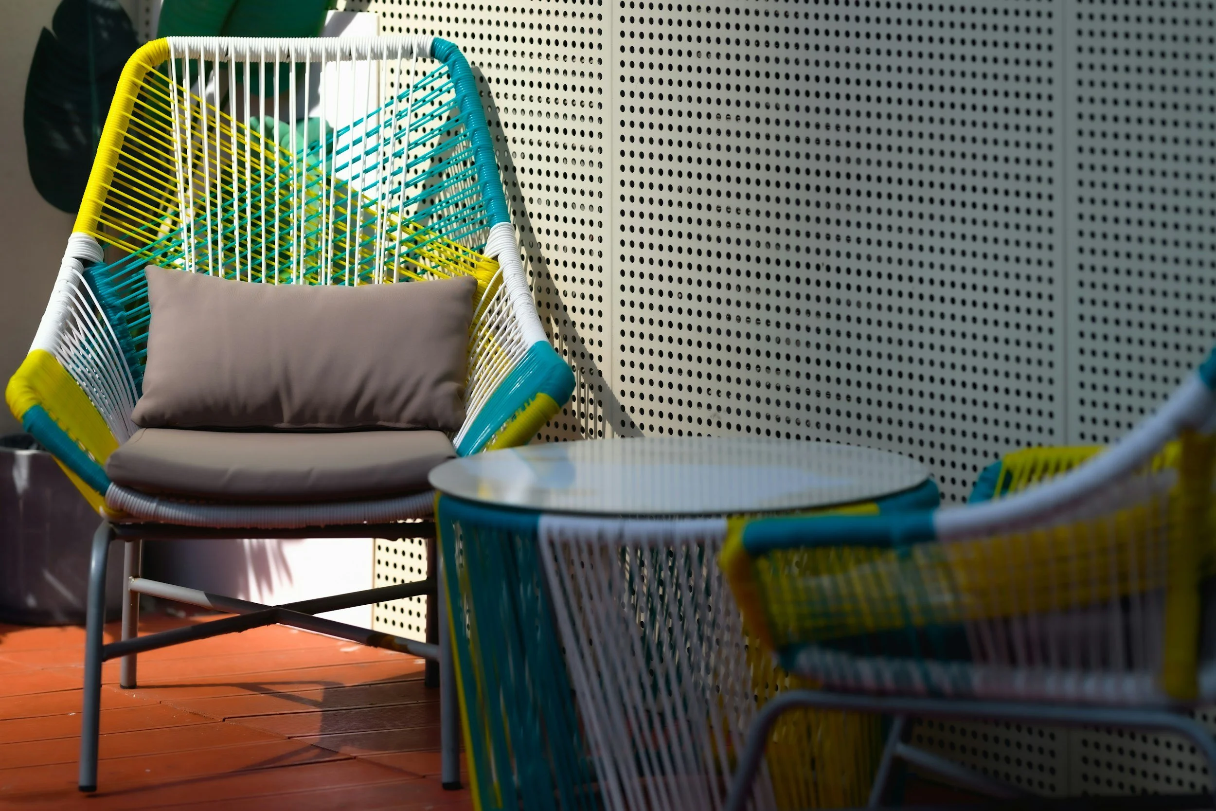 Colorful outdoor chair with beige cushion placed on wooden patio floor, set against a pegboard wall.