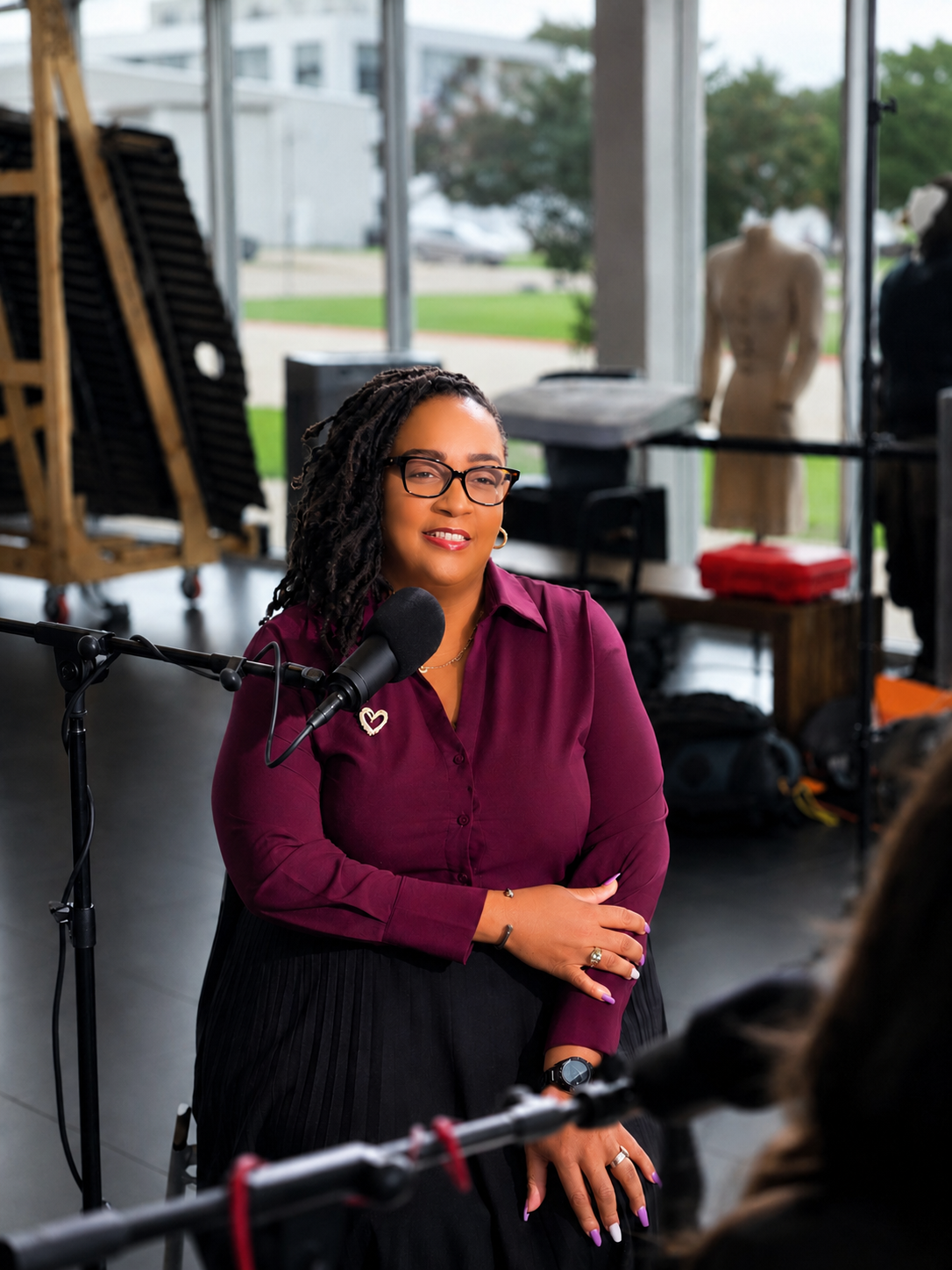 A woman with glasses and dreadlocks is speaking into a microphone during a recording or interview in a room with large windows. There are art mannequins and folding chairs in the background.
