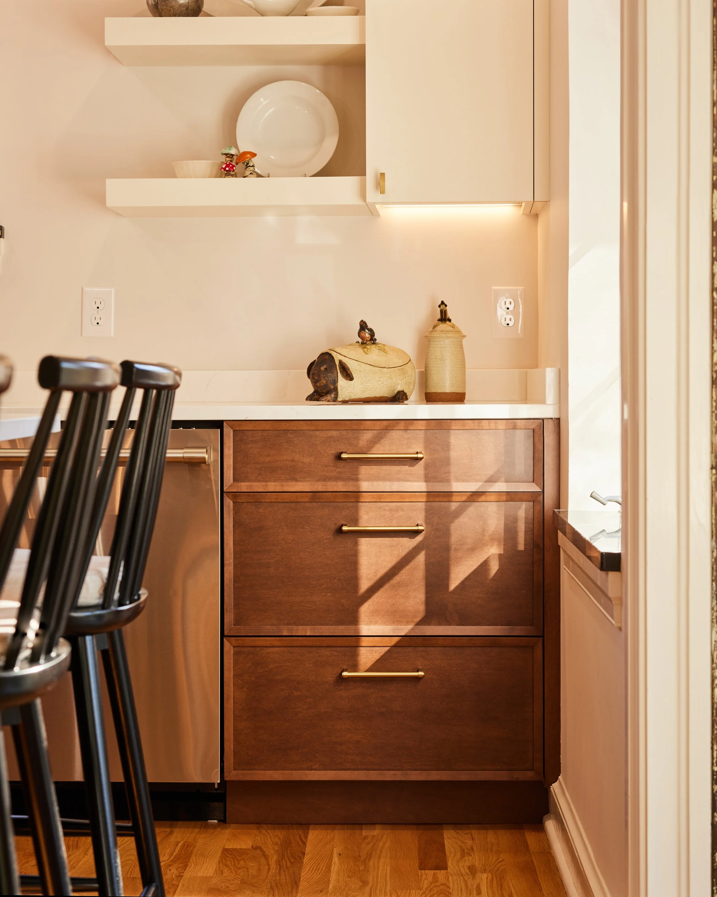 A kitchen corner with wooden cabinets, decorative items, open shelves, and a window letting in sunlight.