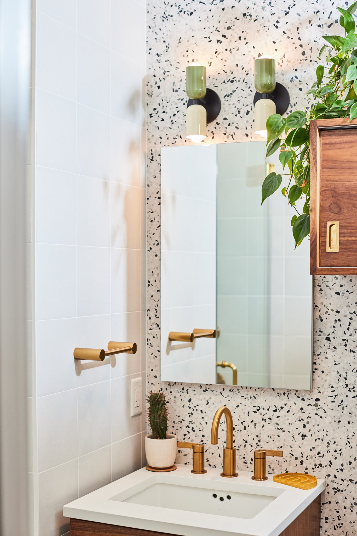 A modern bathroom featuring a mirror, a white sink with brass fixtures, a potted cactus, wall-mounted brass towel holders, stylish light fixtures, and a wooden cabinet with a plant.