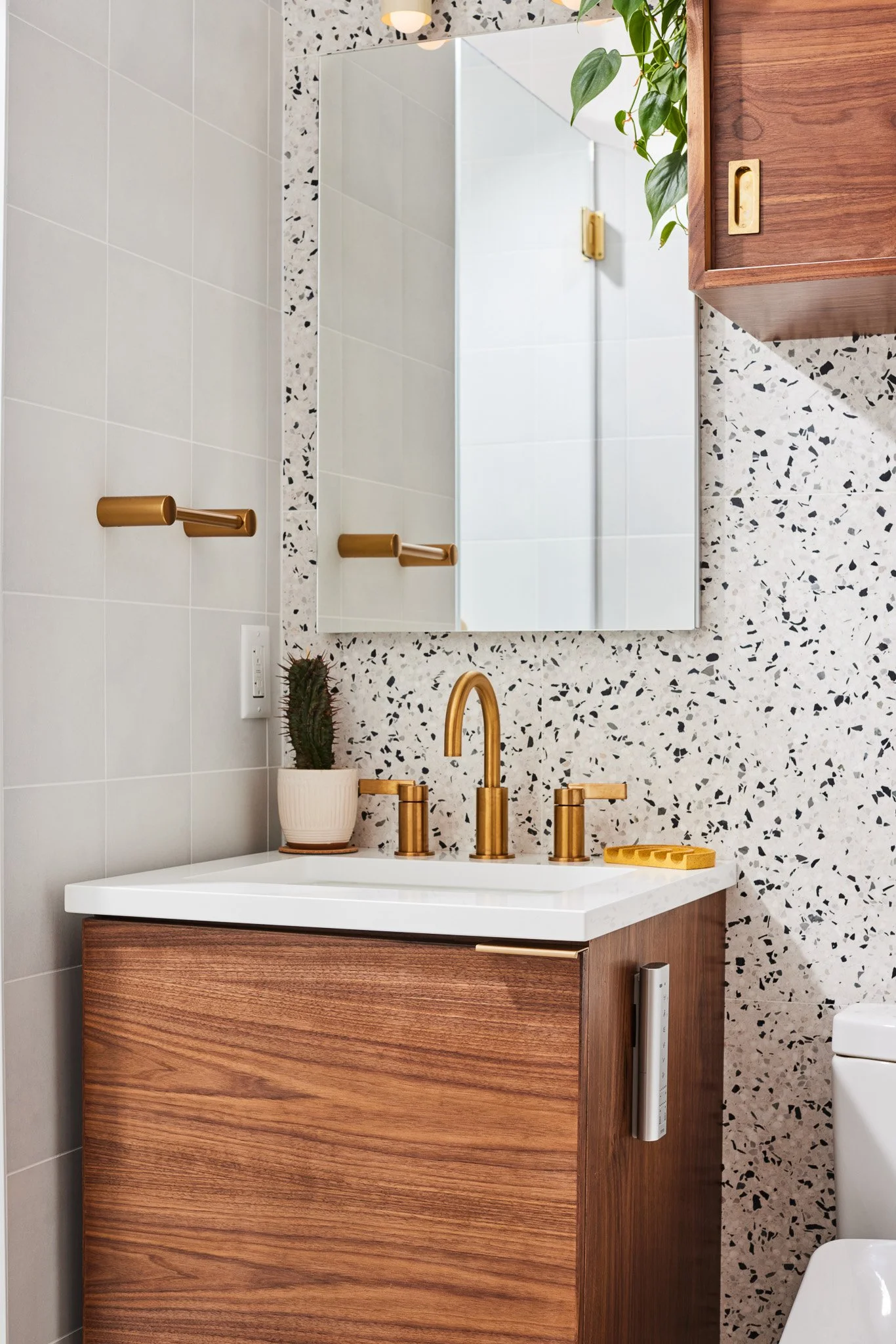 Bathroom vanity with a white sink, brass fixtures, a potted cactus plant, and a wall mirror. There are two gold towel bars and a wood cabinet beside the mirror, with a mirror reflecting white tiled walls.