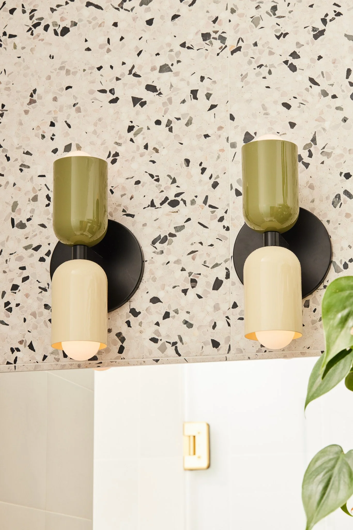 Two wall-mounted light fixtures with black round bases, each with two colored glass shades, one green and one cream, on a terrazzo wall.