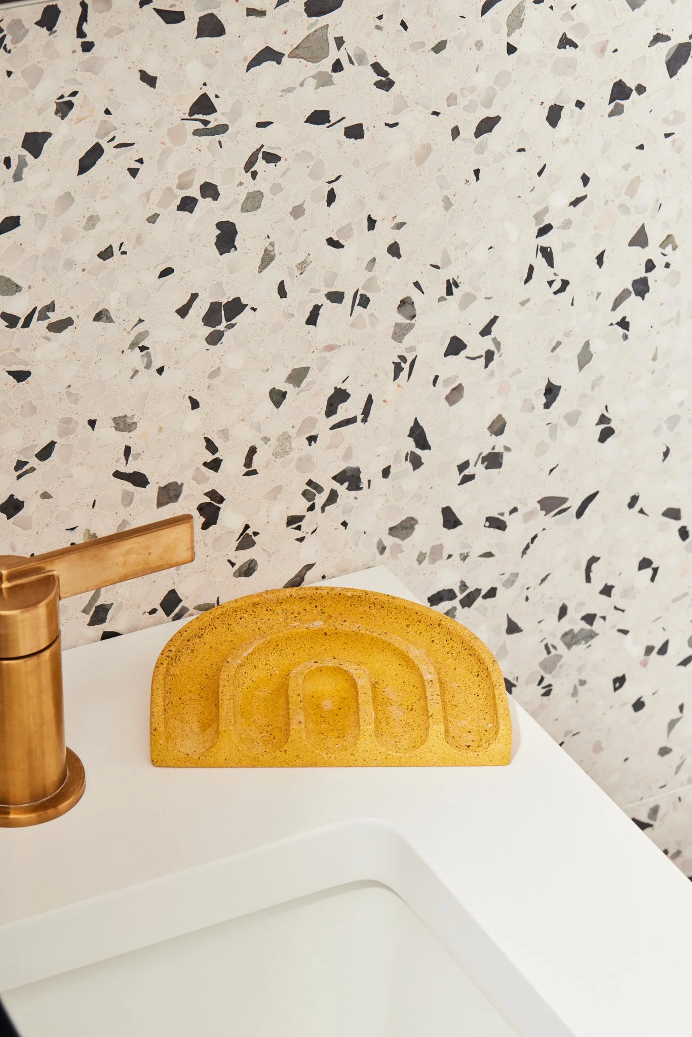 A yellow, textured soap dish with a semi-circular design situated on a white countertop, next to a bronze faucet, with a terrazzo-patterned wall in the background.