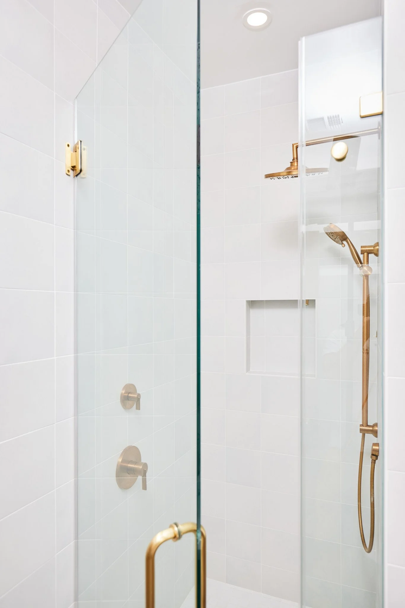 A modern shower with white tiled walls, a glass door with a gold handle, and gold fixtures including a showerhead and controls.