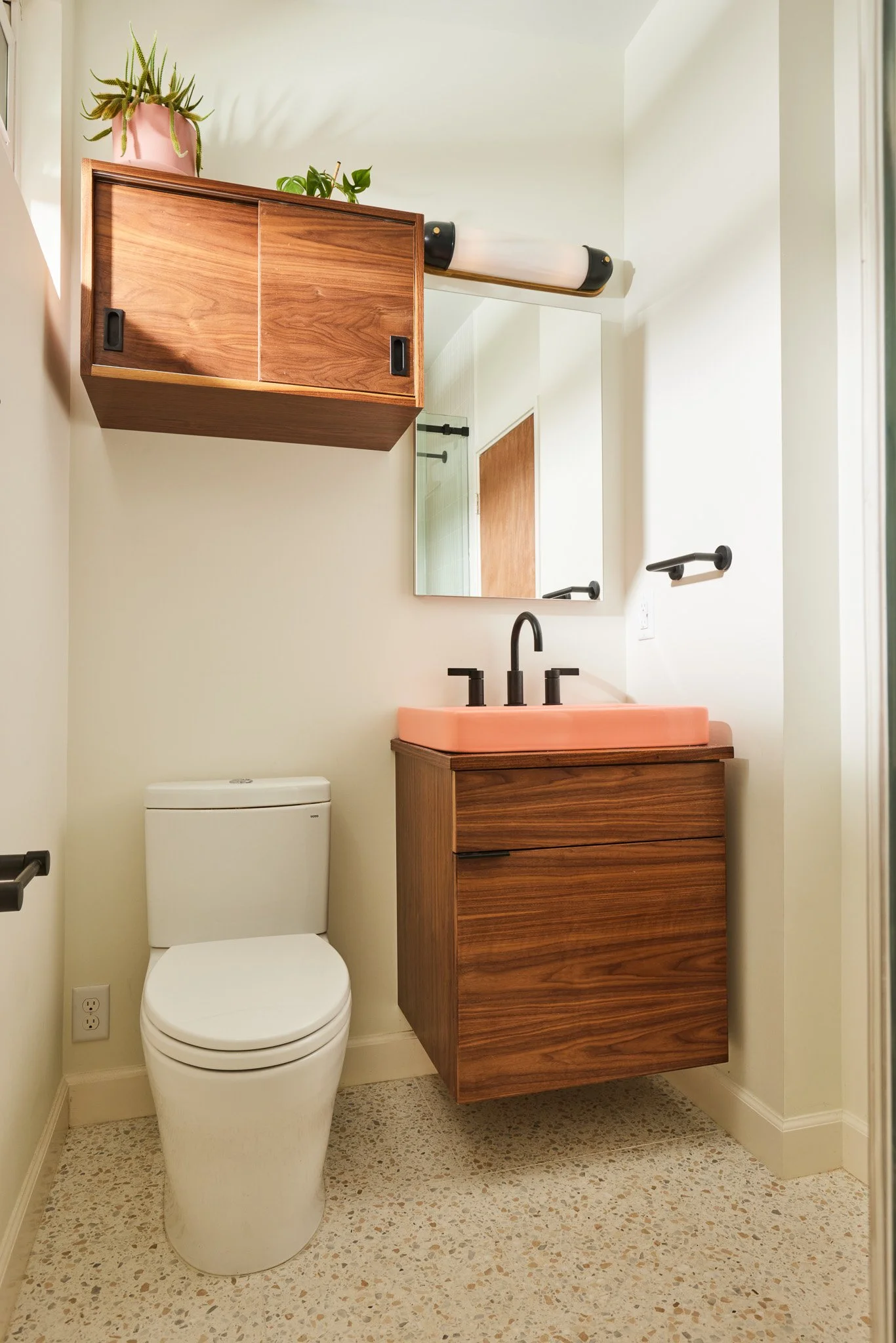 A bathroom with a modern design, featuring a white toilet, a wooden floating vanity with a pink sink, a mirror above the sink, black fixtures, a wall-mounted cabinet, and plants on top of the cabinet.