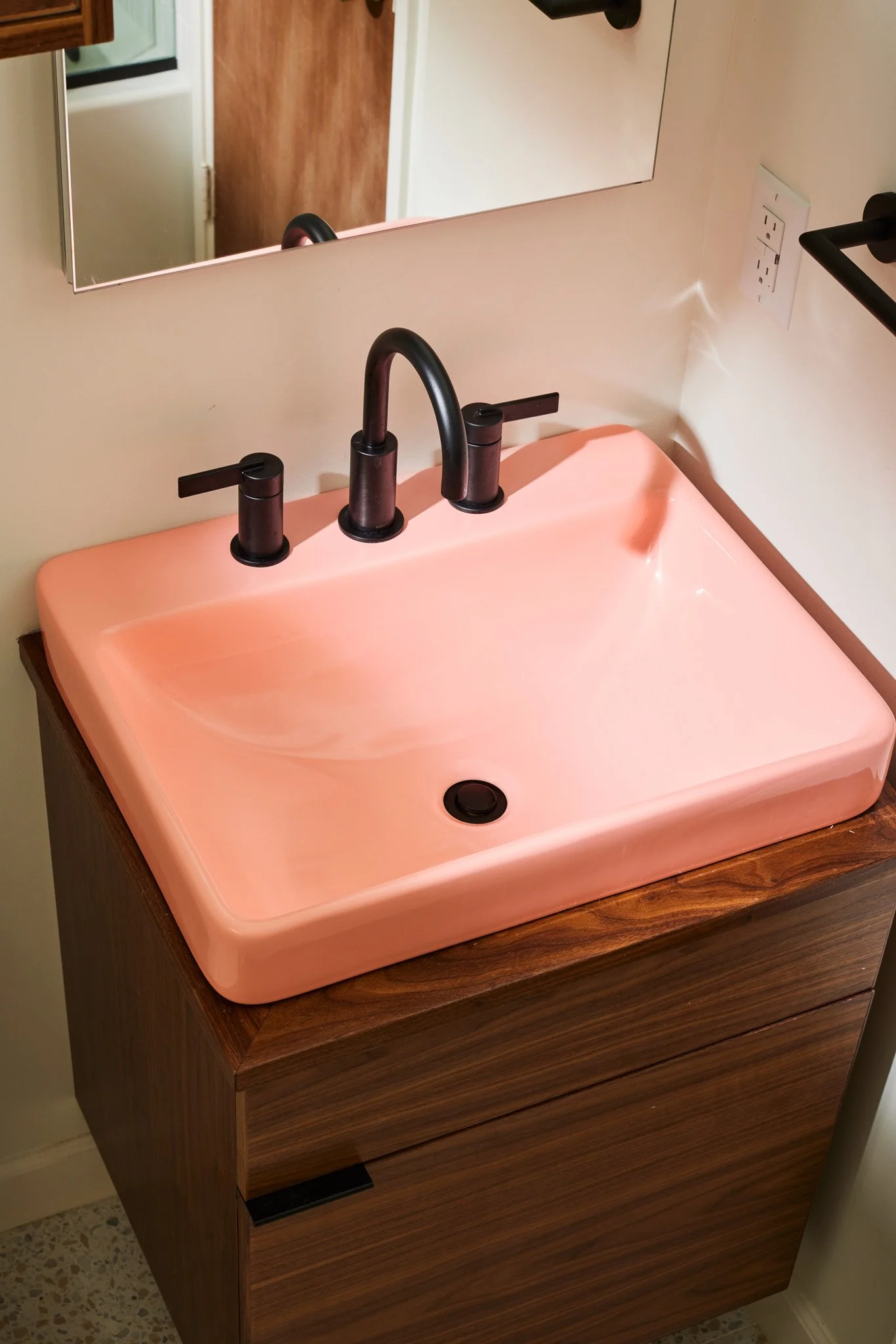 A pink rectangular bathroom sink with black faucet and handles, mounted on a wooden vanity with a mirror above.