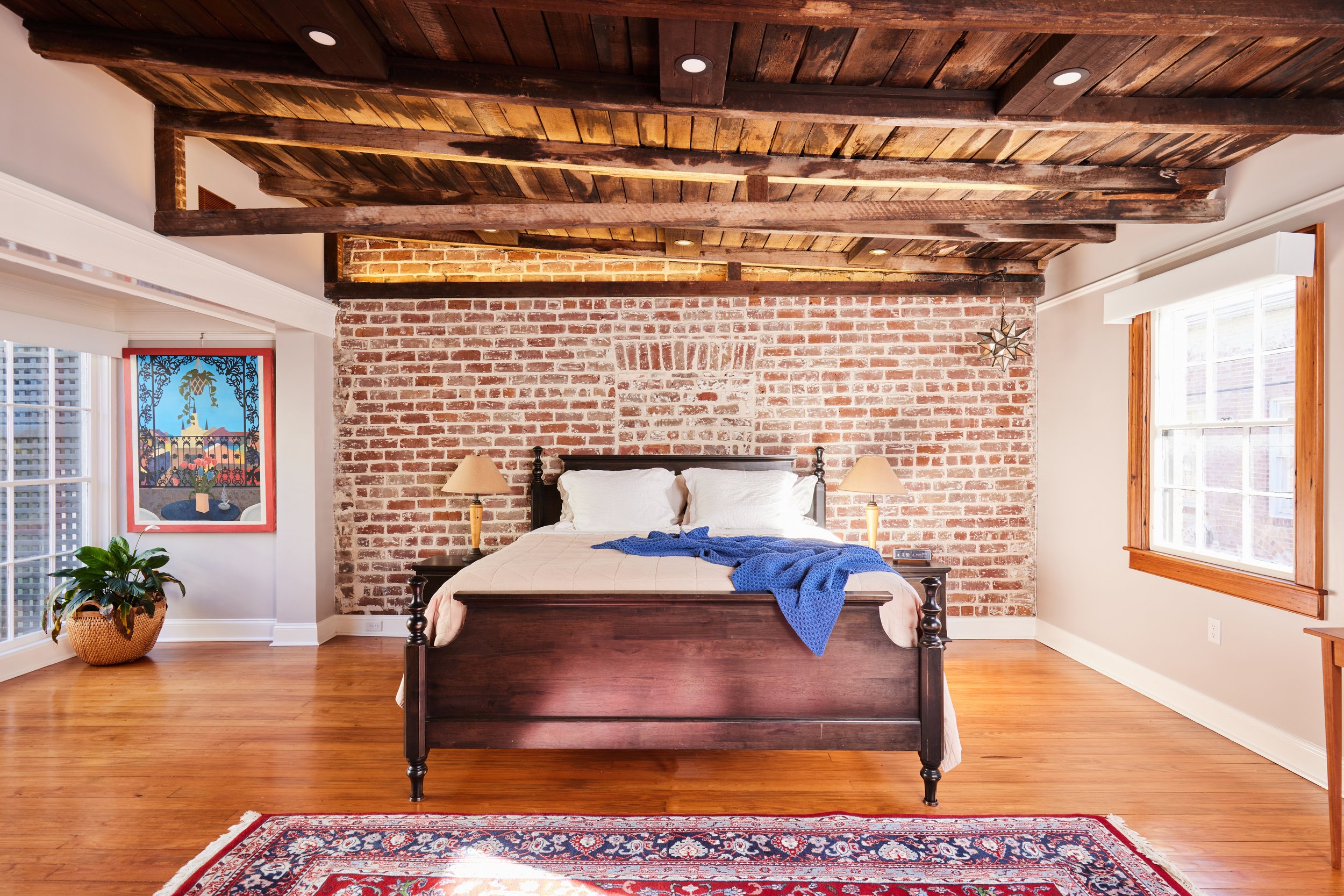 A bedroom with a rustic exposed brick wall, wooden ceiling beams, and hardwood floors. The bed has dark wooden frame, white bedding, and a blue throw blanket. Two small nightstands with lamps are on each side. Large windows with wooden trim let in natural light, and there's a framed colorful artwork and a potted plant in the room.