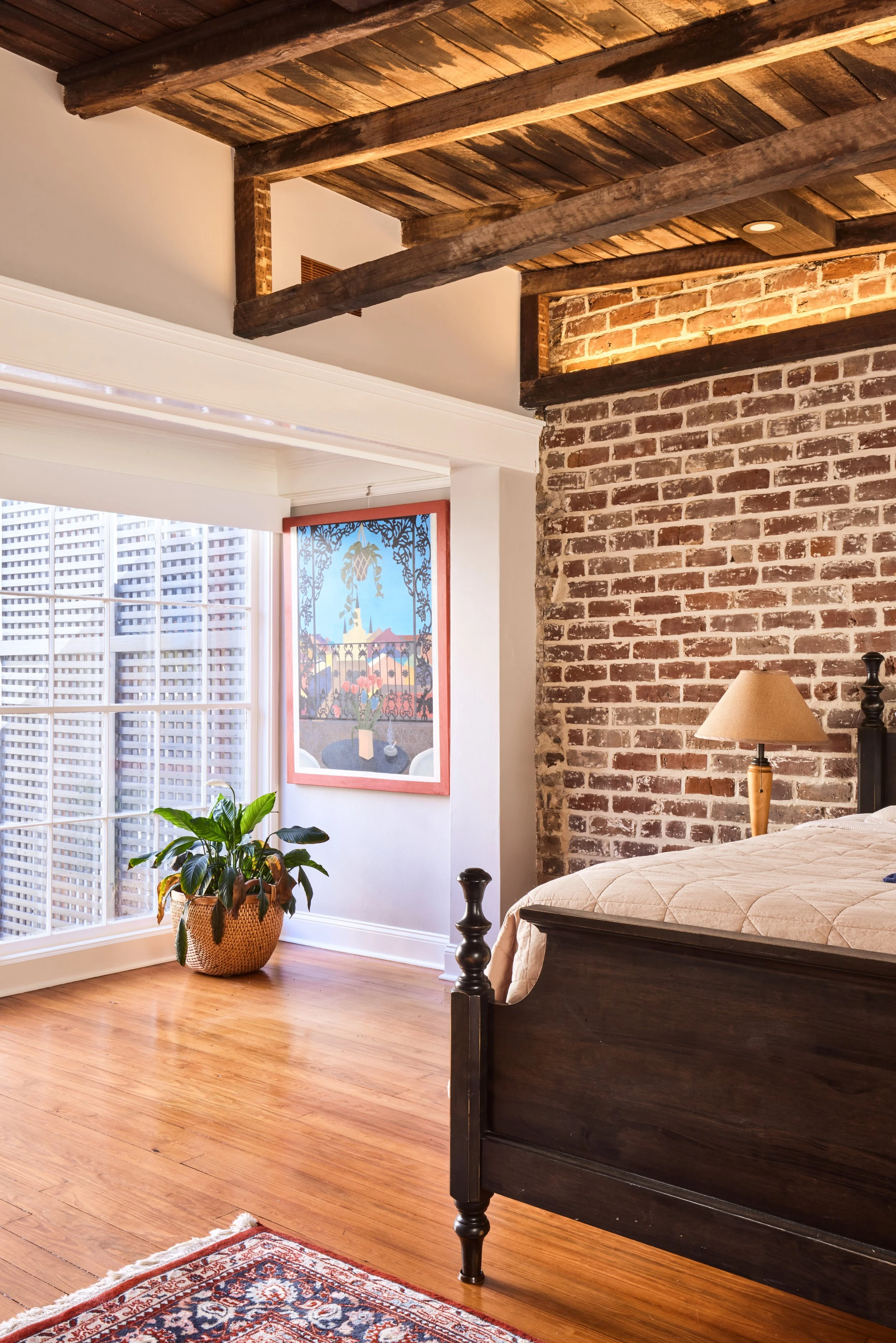 A cozy bedroom with exposed brick wall, wooden ceiling beams, hardwood floors, a dark wood bed frame, a beige quilt, a table lamp, a potted plant, and artwork on the wall near a large window with white blinds.