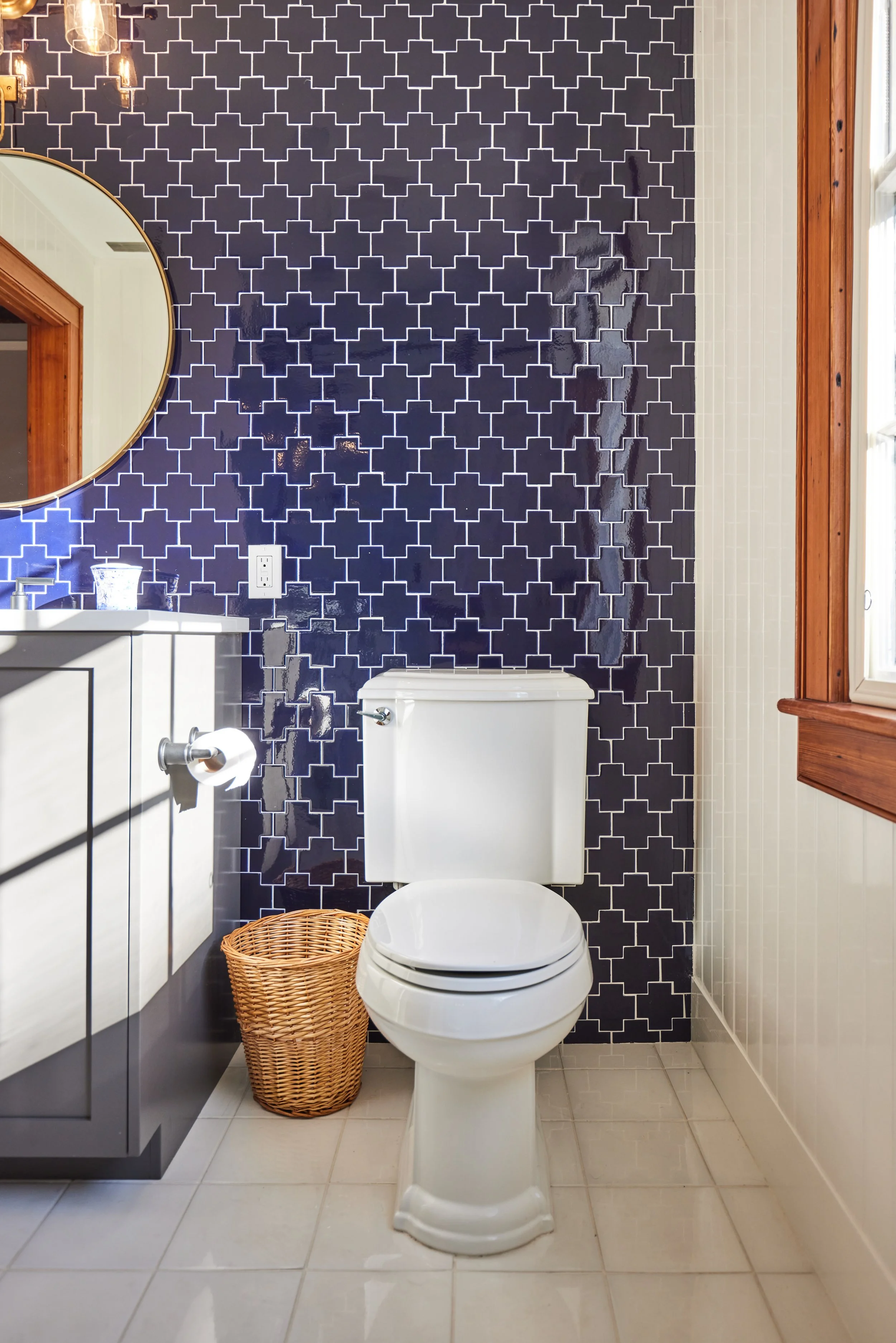 Bathroom with navy blue tiled wall, white toilet, small vanity with a mirror, wicker basket, and a window with wooden trim.