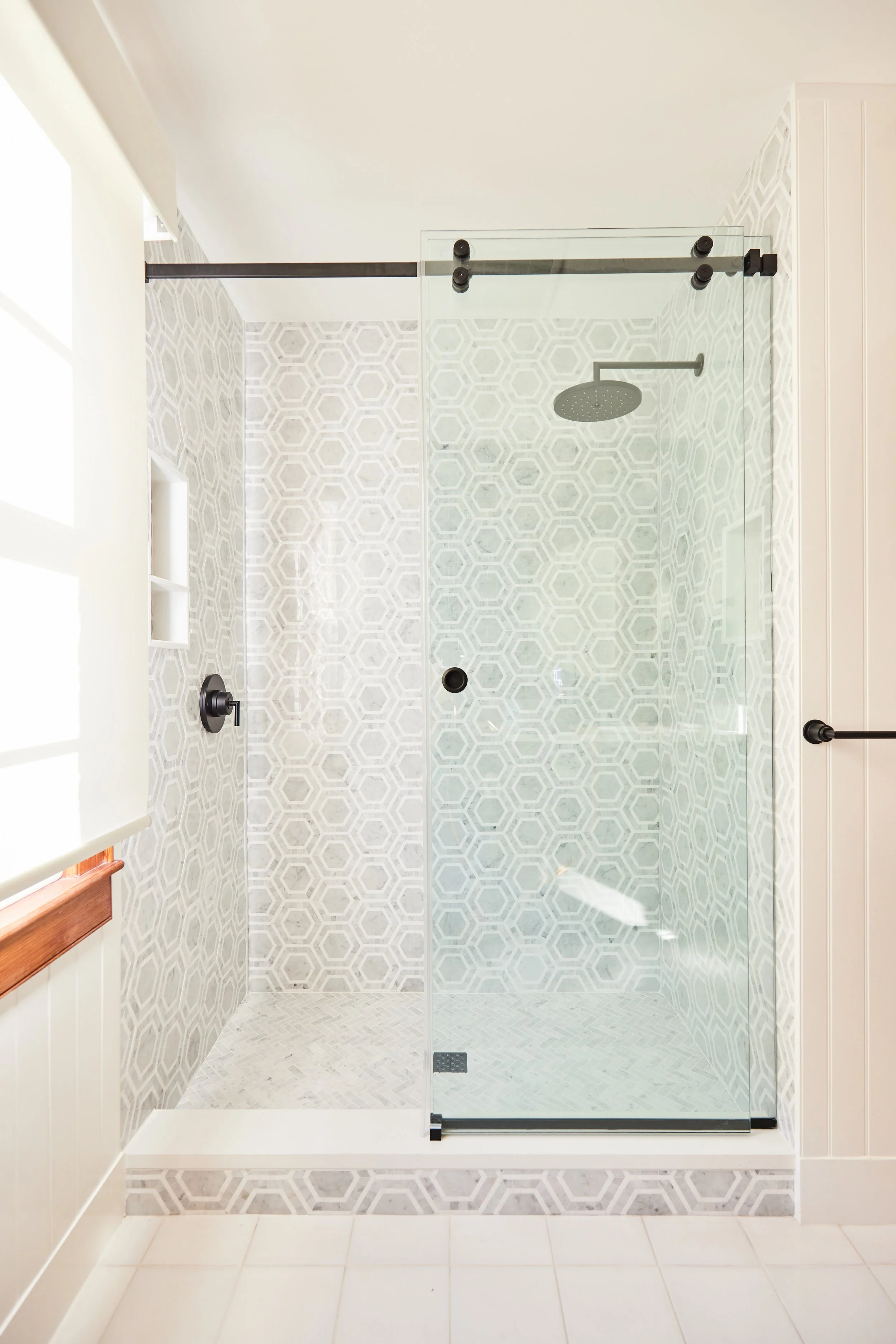 A glass-enclosed walk-in shower with geometric patterned tile walls and floor, a rainfall showerhead, a wall-mounted control, and a built-in shower niche, located in a bright bathroom.